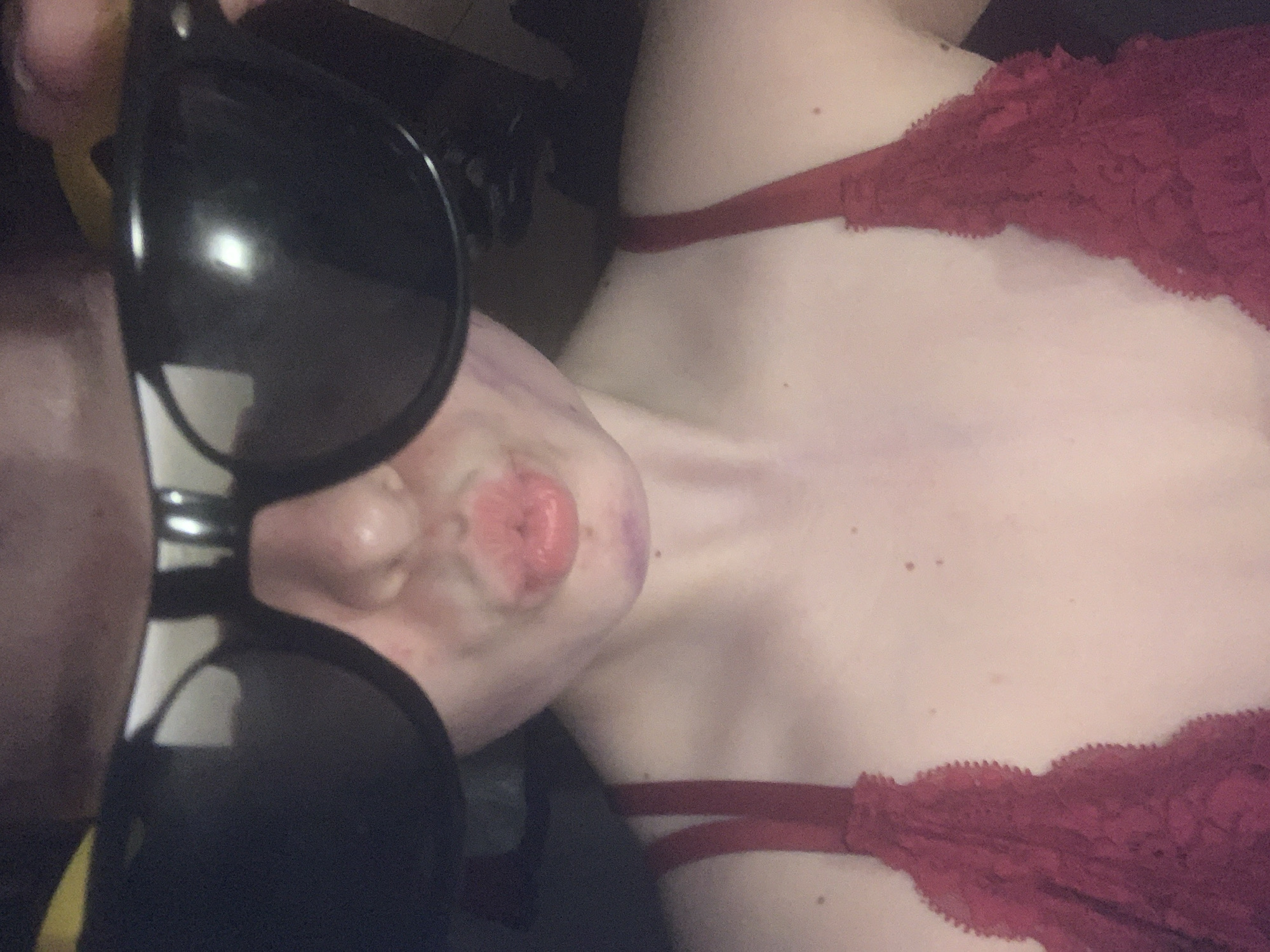 The image presents a close-up, angled view of a person's upper body, taken from a slightly elevated perspective. The subject is a person with fair skin, reclined or lying down, wearing a red lace bra or top. Large, black sunglasses are positioned over their face, obscuring the eyes, with reflections visible on the lenses. The person's mouth is pursed into a "kissing" or "pout" shape. Faint purple or reddish marks are visible on the neck and collarbone area. The background is indistinct, suggesting an indoor setting. There is no discernible activity other than posing for the photograph. No text is visible in the image. The provided location context is Vsevolozhsk, Russia.