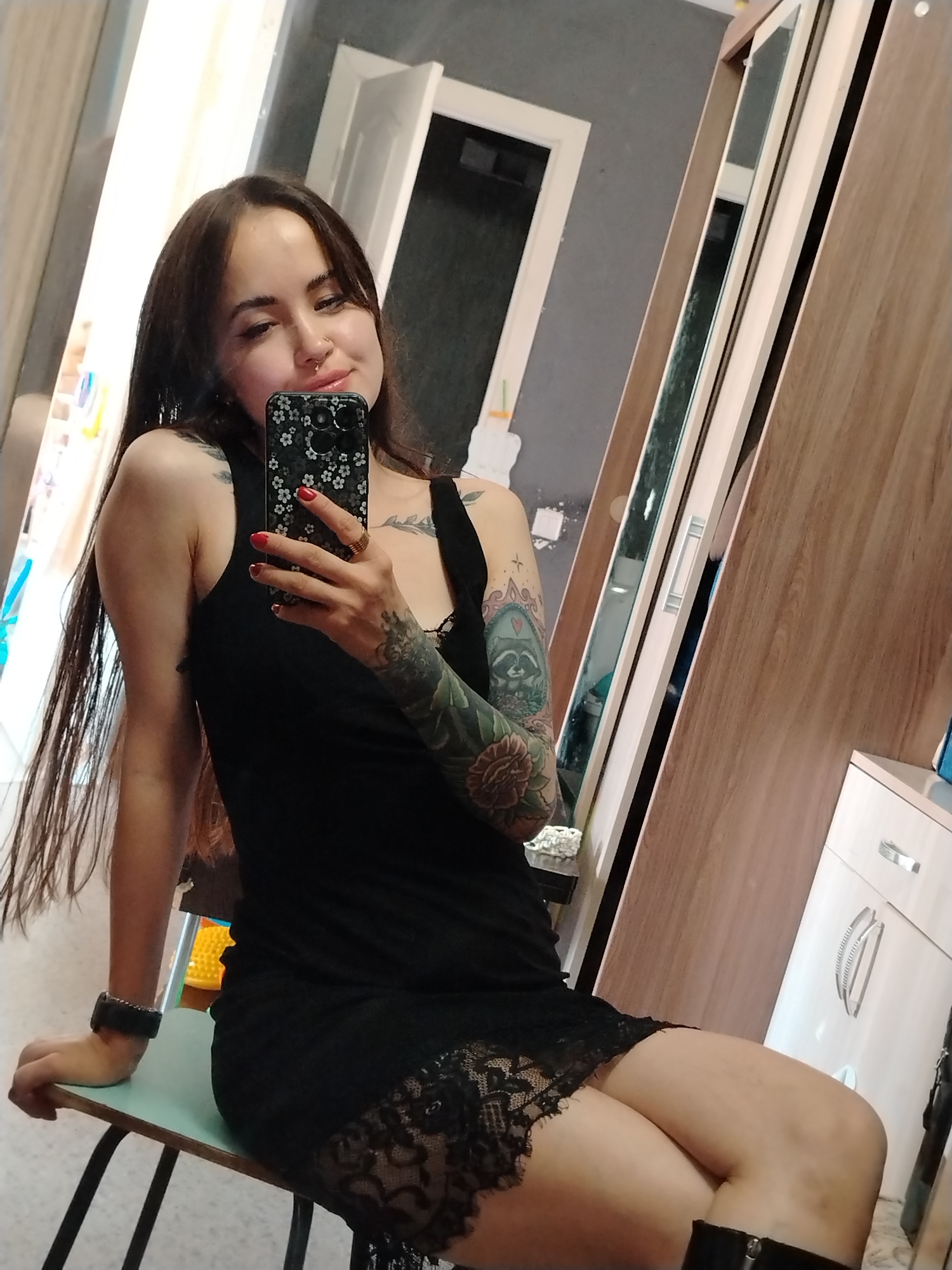 A woman with long dark hair is taking a selfie in what appears to be a bedroom or dressing area. She is wearing a black dress with lace trim and has tattoos on her arm. She is holding a smartphone with a floral case. In the background, there is a wardrobe with mirrored doors and a white cabinet. The environment suggests a domestic setting. The location is identified as Noviy, Russia.