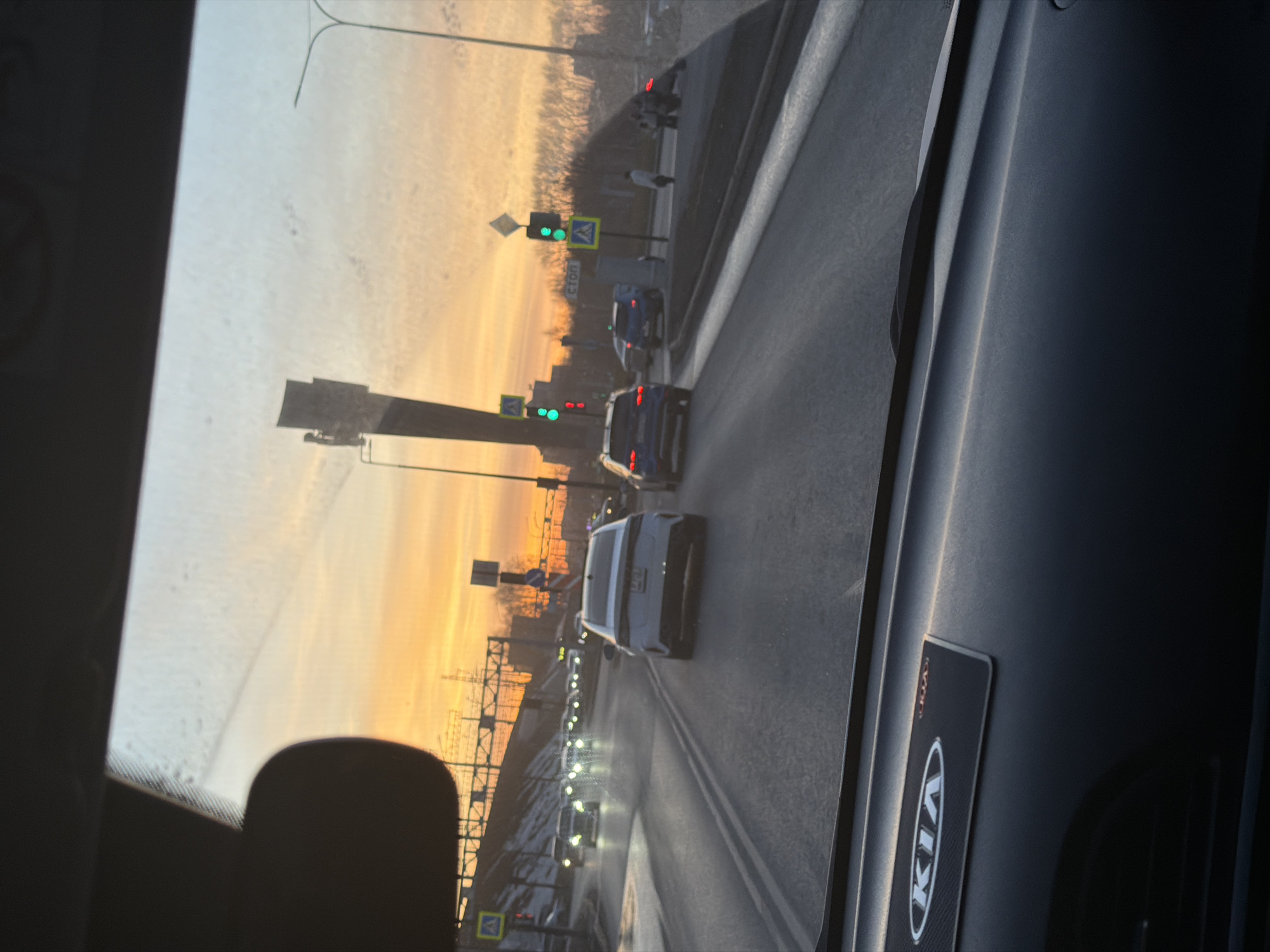 The image shows a view from inside a Kia car, likely the passenger seat. The camera faces forward, capturing a road with multiple lanes of traffic. The cars include a white car and several dark-colored cars. A traffic light shows green signals. The image hints at being taken during sunrise or sunset, with the sky having a strong orange hue. This area is located within Perm, Russia.