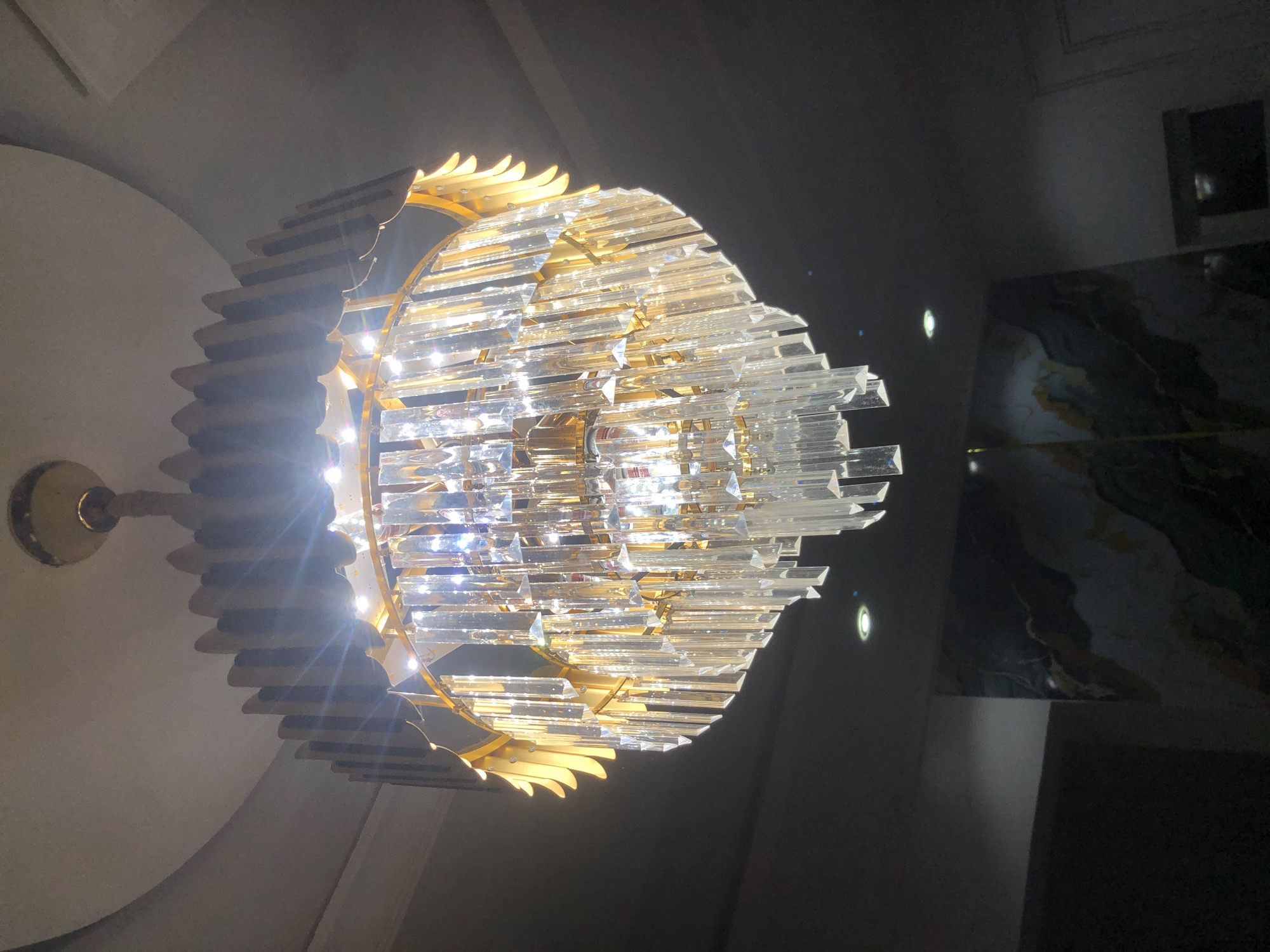 A chandelier hangs from a ceiling in a building. The chandelier has multiple tiers of clear crystal prisms suspended from a gold-colored frame. The prisms are of various lengths and reflect the light. The ceiling is white and appears to have a curved design. The background is dark, with some subtle lighting visible from below, possibly from recessed ceiling lights. There is a wall in the background with a decorative pattern that appears to be marble or a similar material with swirling colors. The overall impression is one of luxury and illumination. The image is taken from a low angle, looking up at the chandelier.