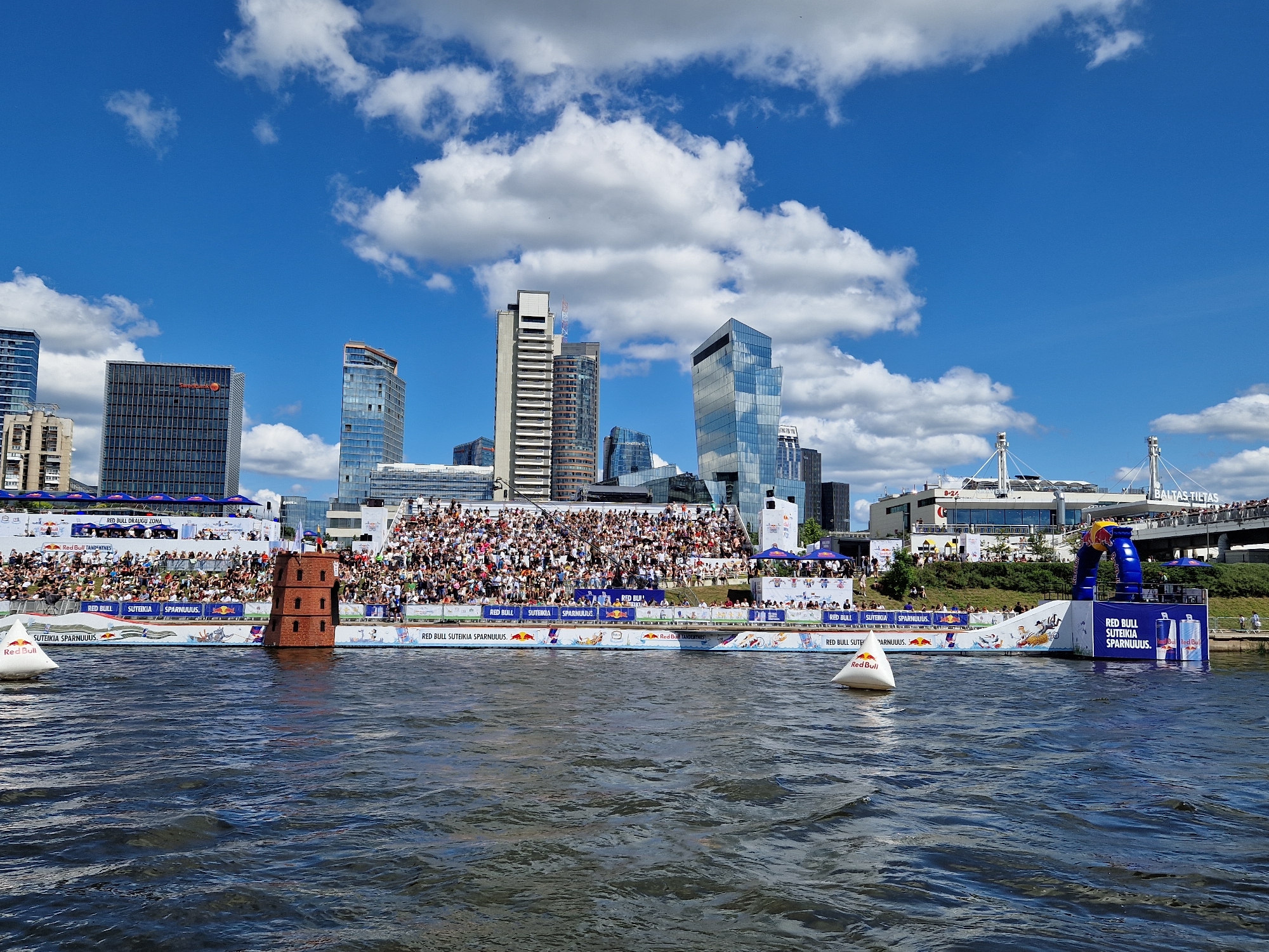 This is a daytime, eye-level, medium-shot photograph of a Red Bull event taking place on the Neris River in Vilnius, Lithuania, with the city's modern skyline in the background. The foreground shows the dark, rippling water of the river. In the midground, a large crowd of spectators is seated in tiered white bleachers that line the riverbank. In front of the bleachers, a red brick structure resembling a three-tiered tower stands in the water. Further to the right, a blue inflatable archway, branded with Red Bull logos, marks the end of a ramp extending into the water, suggesting a stunt or competition area. White buoys, also branded with Red Bull, are visible in the water. The background features a cluster of contemporary glass skyscrapers, characteristic of Vilnius's business district. The sky is a bright blue with scattered white clouds.