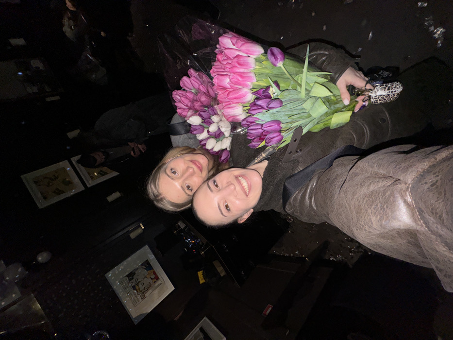 This image depicts two women smiling in a dimly lit indoor establishment in Moscow, Russia.
**People:**
The woman in the foreground, with dark hair pulled back and wearing a dark brown jacket with a shearling-like collar, is holding a smartphone and taking a selfie. She exhibits a broad smile, showing her teeth. Her left hand holds a large bouquet of purple tulips, and a leopard-print keychain is visible attached to her hand.
Next to her, a blonde woman, wearing a grey top, smiles directly at the camera. She holds a large bouquet of pink and white tulips, wrapped in clear plastic.
In the background, at least two additional figures are partially visible in the low light. One male figure, with dark hair tied back, is visible to the right, appearing to look at a mobile phone.
**Subjects/Objects:**
Three substantial bouquets of tulips are prominently featured, wrapped in clear plastic. One bouquet is entirely purple, another is predominantly pink with some lighter pink/white flowers, and a third, held by the blonde woman, is a mix of pink and white. The aforementioned leopard-print keychain is attached to the foreground woman's hand.
**Environment:**
The setting is an indoor space characterized by dark walls and low, atmospheric lighting, suggesting a bar or lounge environment. Framed artwork adorns the dark walls. A prominent piece on the left features a pop-art style image resembling Roy Lichtenstein's "Drowning Girl," depicting a blonde woman crying with a speech bubble. Other framed pieces are visible further back but are indistinct due to the darkness. Small, reflective elements are visible on the ceiling or upper wall. A dark counter or furniture piece is also present in the background. The overall illumination creates deep shadows and highlights subjects in the immediate foreground.