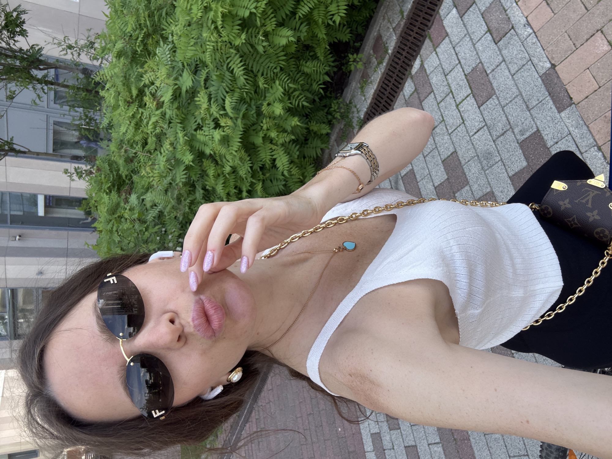 This full-frame selfie captures a young woman outdoors in what appears to be an urban setting in Moscow, Russia. The image is taken at an oblique angle, with the woman looking directly at the camera.
The central subject is a woman with long, dark brown hair, wearing large, dark sunglasses with gold frames. The lenses of the sunglasses feature the "FF" logo, indicating they are Fendi brand. She wears white earbuds. Her lips are playfully puckered in a "kissing" pose, and her left hand is raised near her face, showcasing light pink, glittery nail polish.
Her outfit includes a white ribbed tank top. She is accessorized with multiple pieces of jewelry: a delicate gold chain necklace with a small turquoise heart-shaped pendant, another thicker gold-colored chain necklace, a gold bracelet with a small charm, and a silver and gold-toned watch on her left wrist. A small, brown Louis Vuitton bag, identifiable by its classic "LV" monogram and gold chain strap, is visible in the lower right corner of the frame.
The background features a lush wall of green fern-like plants to the left, behind which a modern building with large, rectangular windows can be seen. To the right, a paved ground consisting of alternating light grey and reddish-brown rectangular tiles is visible, alongside a metal drainage grate. The lighting suggests it is daytime, likely during pleasant weather. The overall mood conveyed by the woman's pose and expression is playful and confident.