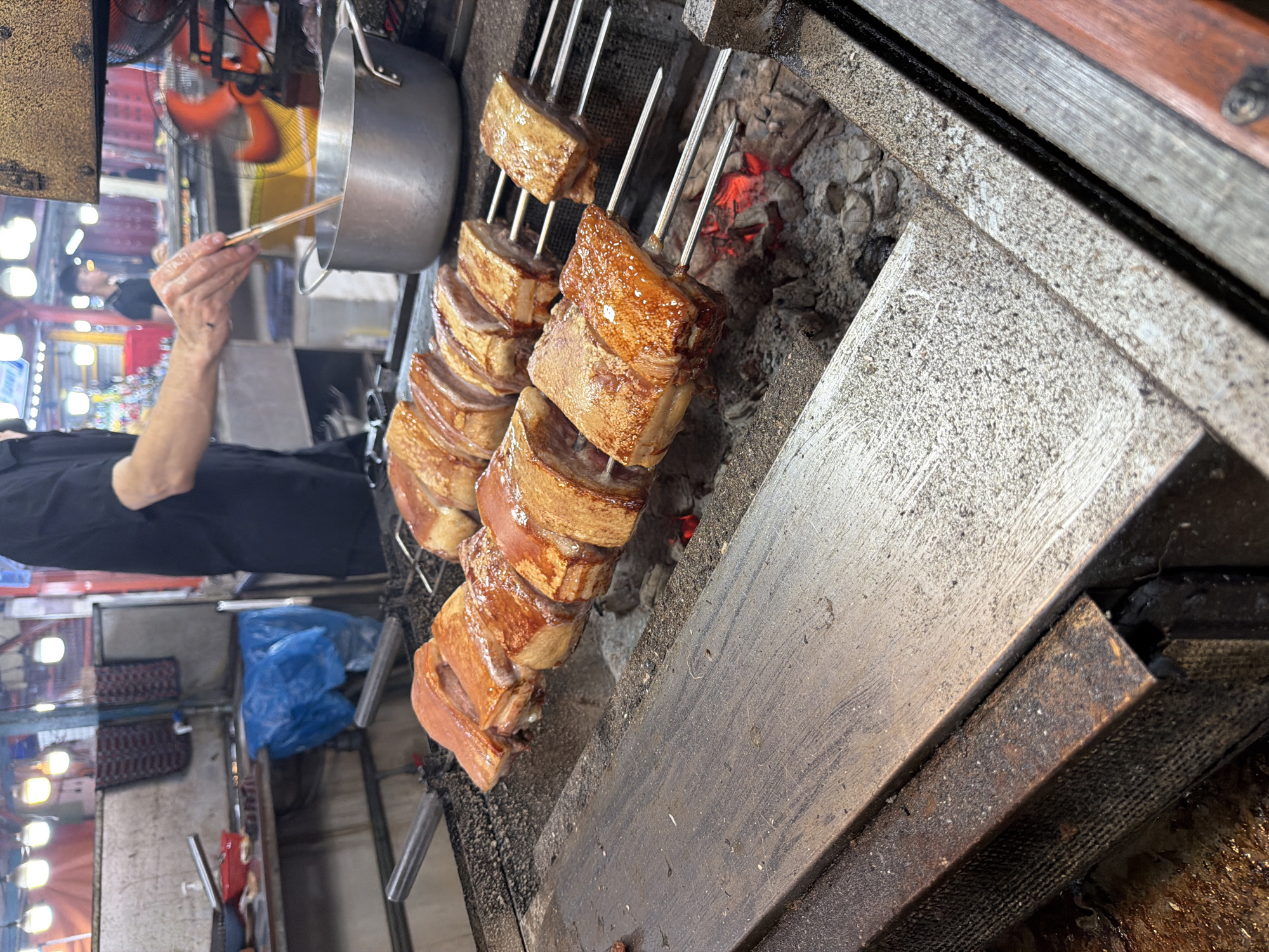 The image depicts a street food stall in Da Nang, Vietnam, where a person is preparing roasted pork. The stall features a charcoal grill with several skewers of pork belly cooking over hot embers. The pork has a crispy, browned exterior with visible layers of fat and meat. In the background, there are market stalls with lights and various food preparation items, including a large metal pot and a blue plastic bag. The scene is bustling and appears to be part of an outdoor market or food street.