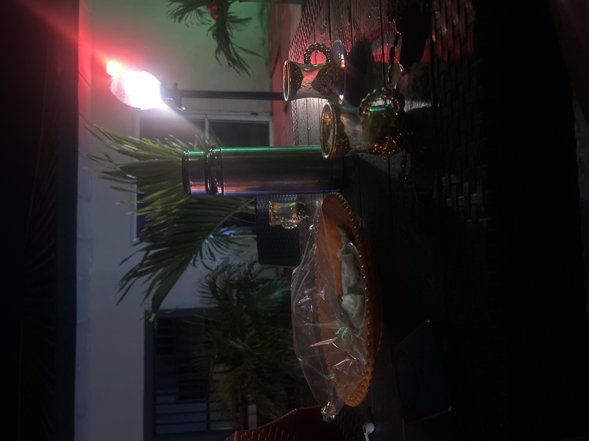 The image depicts a dimly lit outdoor scene, possibly a patio or courtyard, at night. In the foreground, a dark, textured surface, likely a table, is visible. On the table, there are several objects: an orange, decorative plate covered in plastic wrap, containing what appears to be sliced fruit or pastries. Next to it, there is a tall, metallic thermos. Two small, ornate glasses, one with a gold rim, are also present, with one appearing to be partially filled with a light-colored liquid and ice. Further to the right, a single, ornate cup with a handle sits on a saucer. A bright light source, possibly a decorative lamp or torch, casts a vibrant red and green glow on the surroundings, illuminating some of the tropical-looking plants in the background. The overall ambiance suggests a relaxed setting for drinks and perhaps a snack. The location is identified as Masaka, Nigeria.