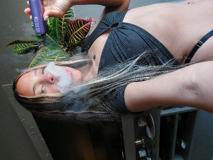 A female individual is depicted indoors, exhaling a cloud of white vapor from her mouth while holding a purple, cylindrical electronic device, consistent with a vape pen or portable vaporizer, in her left hand. She has long, multi-toned hair, featuring dark strands with blonde highlights and some braided sections. She is wearing a black ribbed crop top with a central ring detail, exposing her midriff, and a black strap, likely from undergarments, is visible below her waist. A navel piercing is present. In the background, to the right, a large houseplant with prominent green and red-veined leaves is visible. To the left, a dark flat-screen television is mounted above a shelving unit containing various household items, including candles and other decorative objects. The specific location within Buenos Aires, Argentina, cannot be determined from the visual information provided in the image.