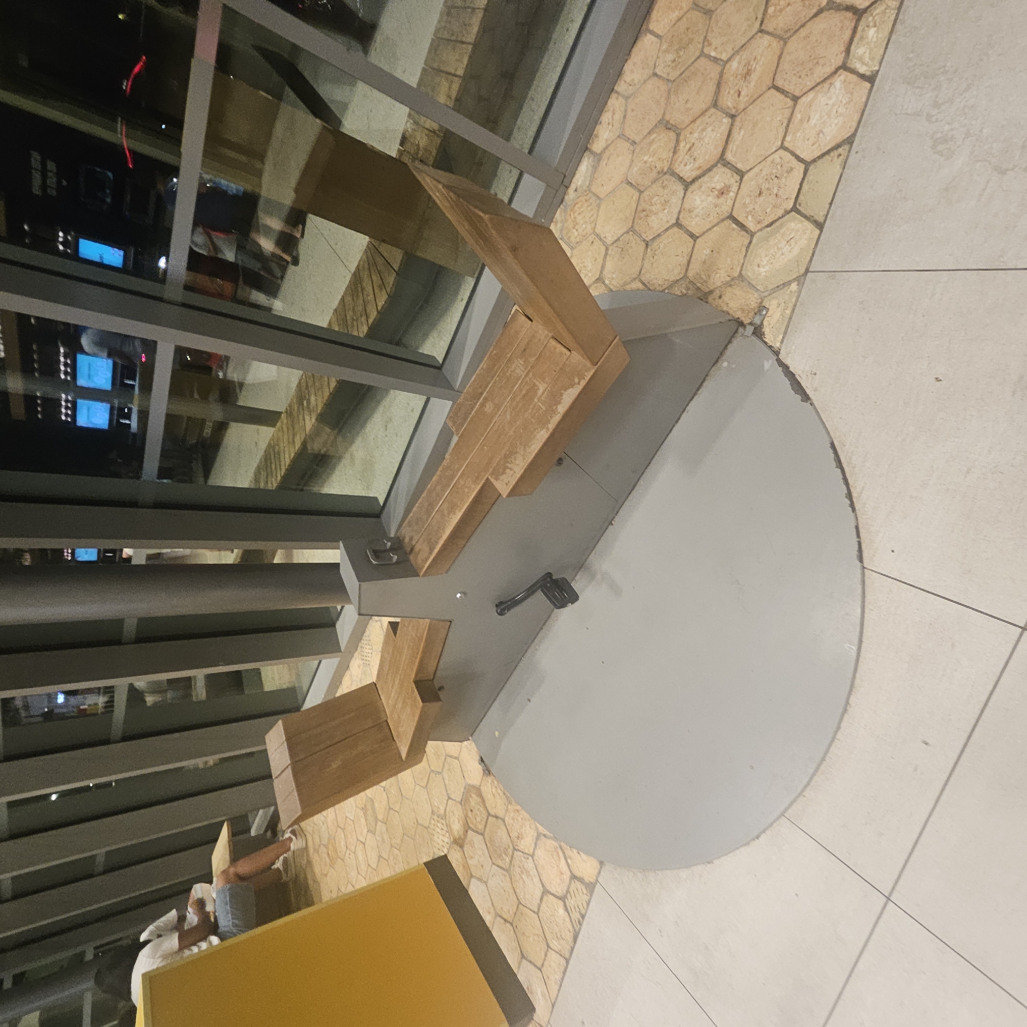 The image shows an exercise machine with wooden seats and a grey base, located on a tiled floor. The machine has pedals. In the background, there are large windows showing a street scene at night, with lights and reflections of people and screens. The overall environment appears to be a public indoor space, possibly a restaurant or waiting area. The location is Lançon-Provence, France.
