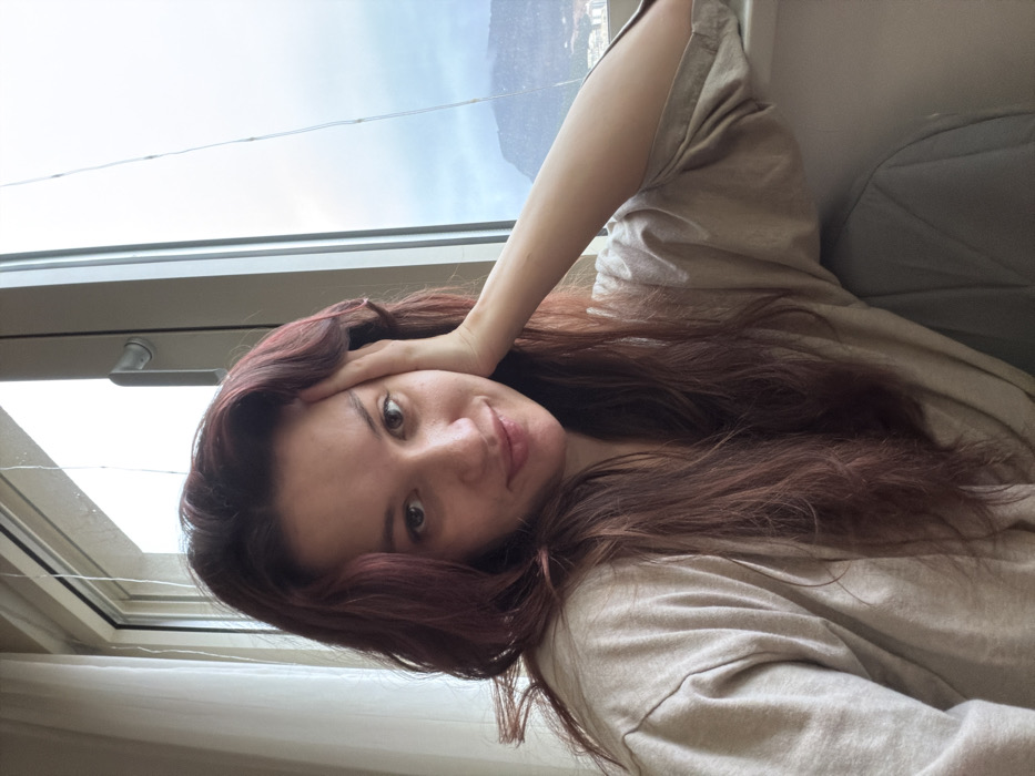 A woman with auburn hair rests her head on her hand, looking at the camera. She wears a light beige top. A window is behind her to the left, with visible clouds and a mountain in the distance. There is a glimpse of a white curtain at the bottom of the image and a gray pillow or cushion towards the top right.