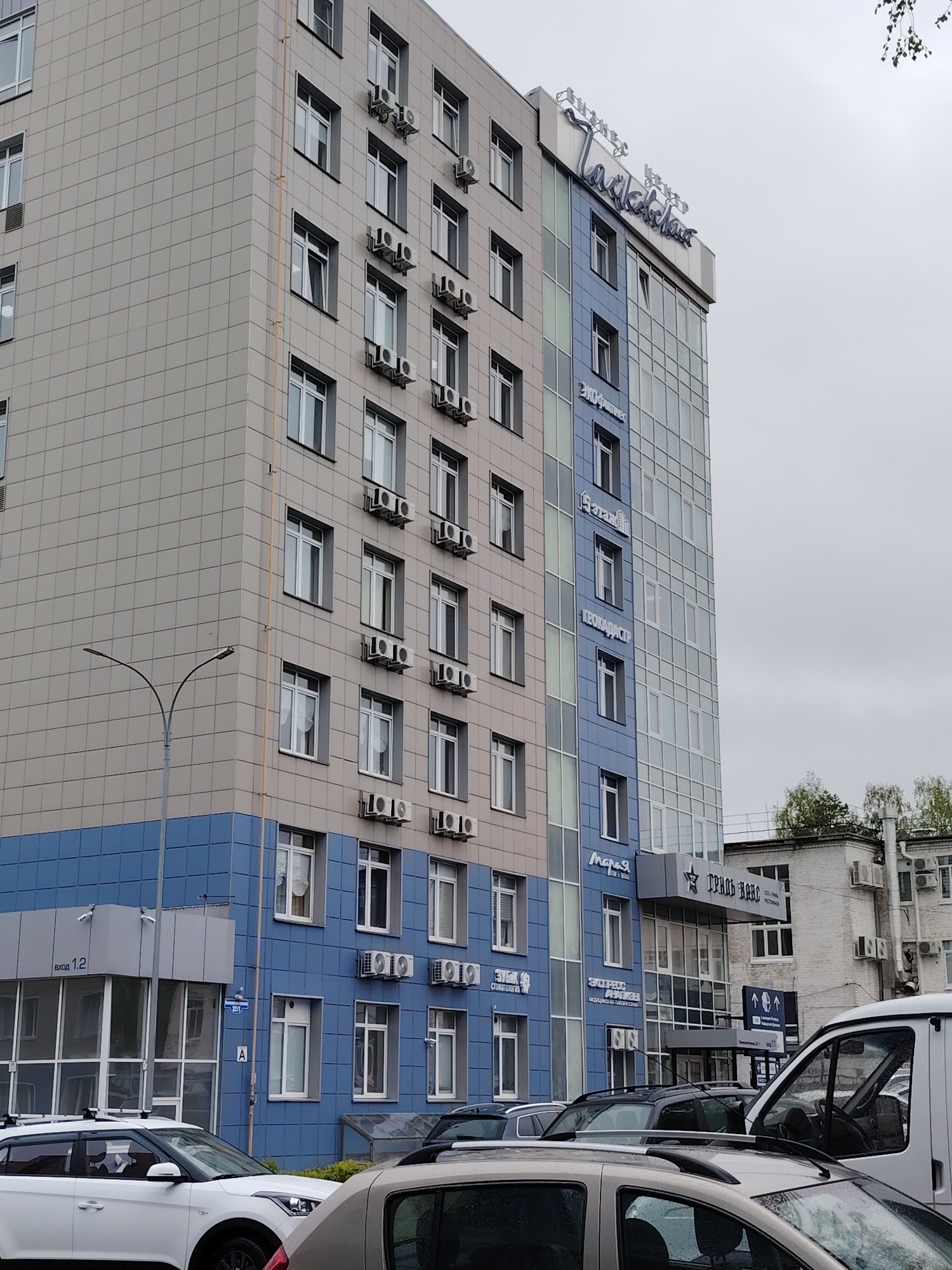 A street scene in Chaikovsky, Russia, depicts a multi-story building with a gray and blue facade. The building features numerous windows and external air conditioning units. Signage on the building indicates various businesses, including "Талисман" at the top. In front of the building, several cars are parked, including a white SUV and a white van. A street lamp is visible on the left side of the image. The sky is overcast. Another building, white with signage that partially reads "Гриль Хаус," is visible in the background.