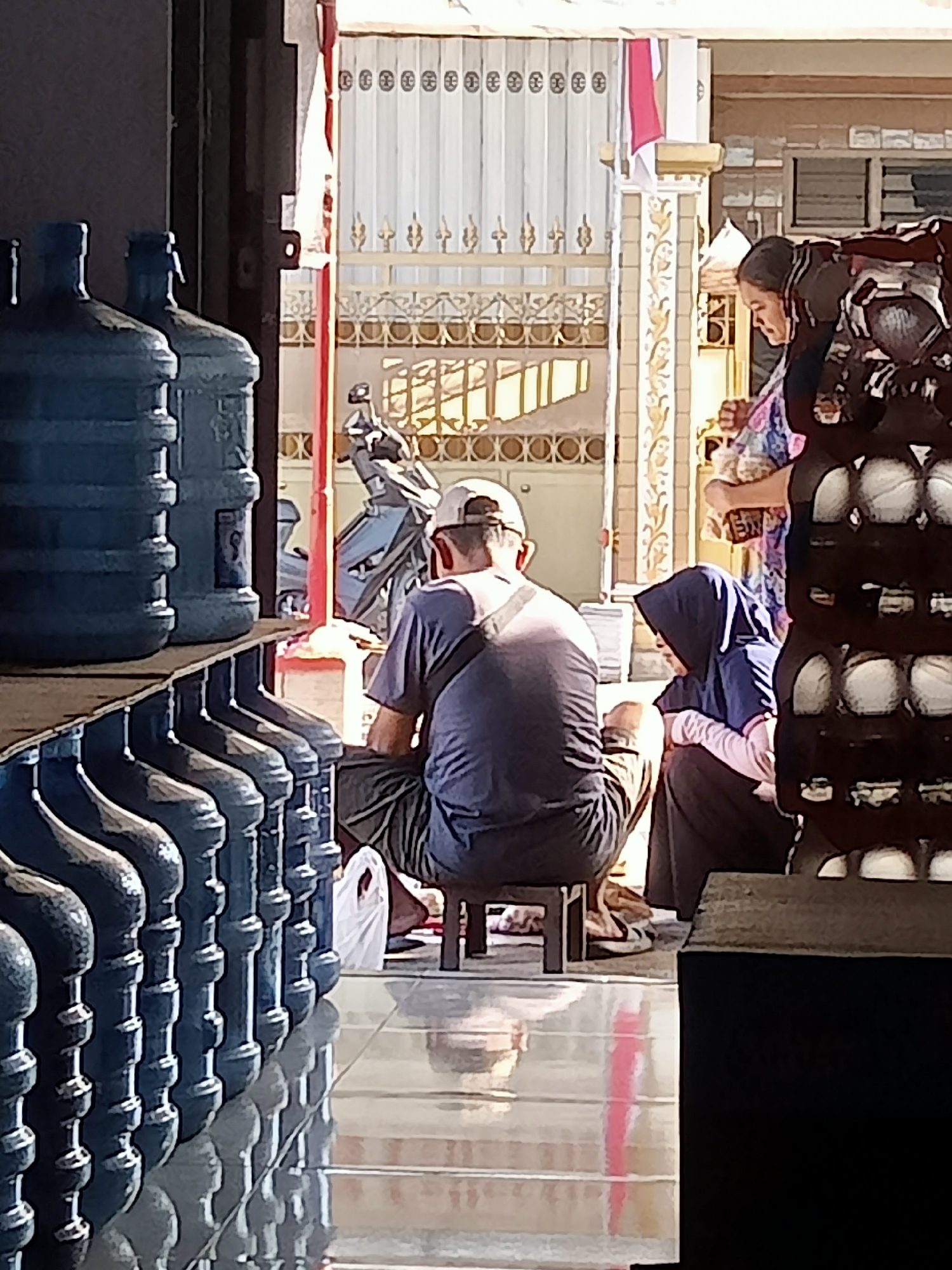 The image depicts a scene in Simo, Indonesia, likely outside a shop or establishment. In the foreground, to the left, is a row of large blue plastic water jugs stacked on a shelf. Further to the right, a man wearing a dark t-shirt, shorts, and a baseball cap is sitting on a small stool. He is facing away from the camera and appears to be looking down. To the right of the man, a woman wearing a blue hijab and dark clothing is sitting on the ground. She is looking down and appears to be interacting with the man or an object on the ground. Behind them, a motorcycle is parked, and there is a decorative gate or fence with an Indonesian flag hanging from it. The overall environment suggests a warm, sunny day.