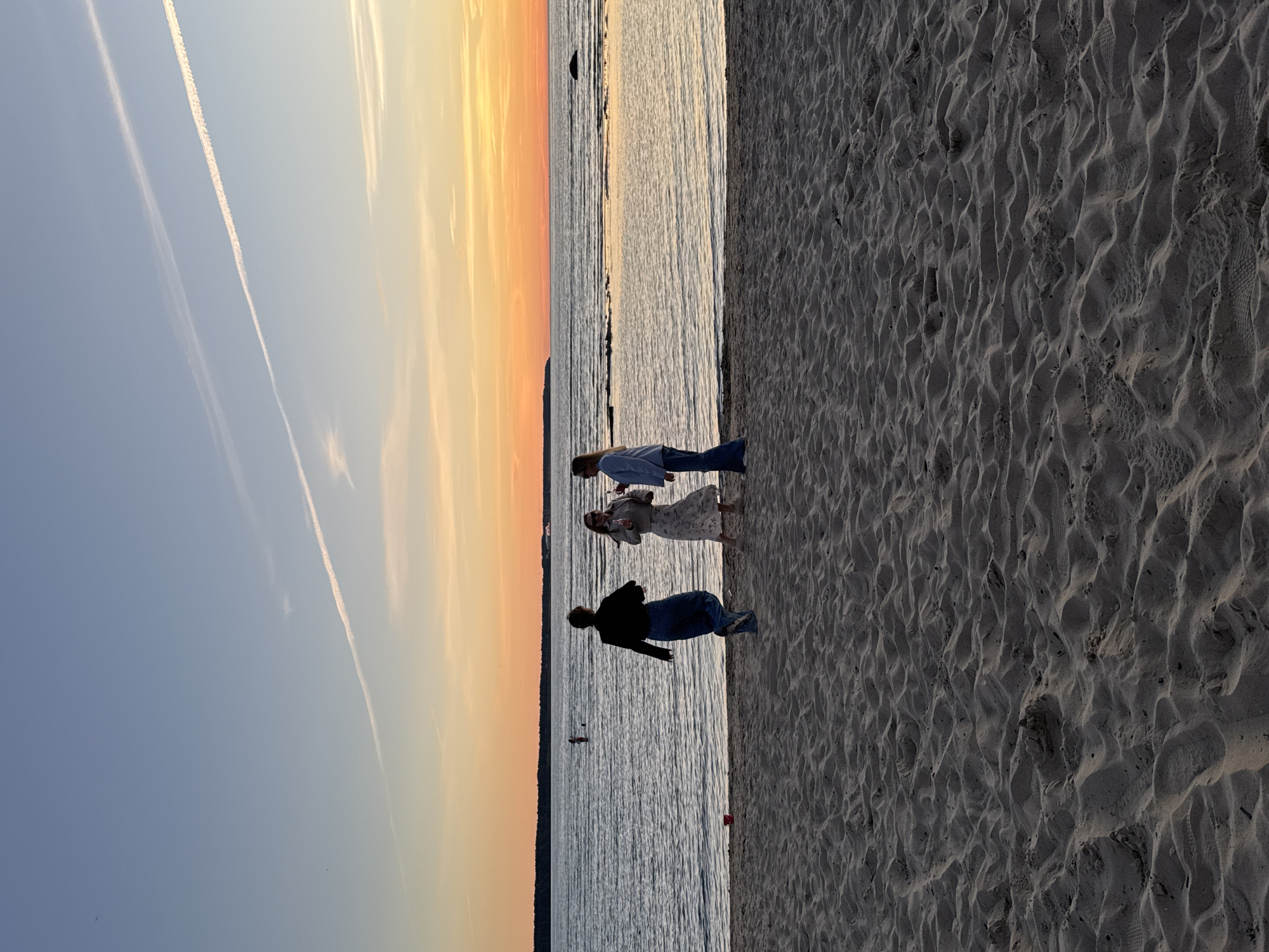 This image depicts a sunset scene at a beach in Tallinn, Estonia. In the foreground, there is a sandy beach with subtle ripples and footprints. Three individuals are visible on the beach, standing near the water's edge. One person, wearing a dark jacket and blue jeans, is facing away from the camera, seemingly walking or dancing. To their right, another person in a light-colored dress and a light jacket is holding a drink and talking to a third person in a blue shirt and jeans. The water is calm, reflecting the colors of the sky. In the distance, across the water, there is a treeline and the faint outline of buildings or structures. The sky displays a gradient of colors, from a pale blue at the top to warm oranges and yellows near the horizon, with two distinct contrails from airplanes streaking across the sky. A small red object is visible on the sand to the left.