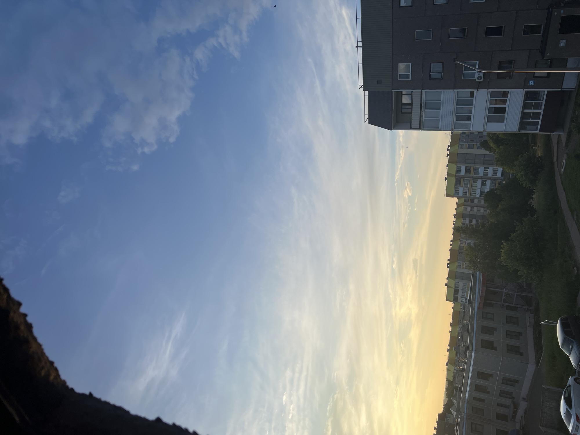 This is a medium-shot, eye-level, vertical, daytime, exterior photograph of a residential area in Markovsky, Russia. The image depicts apartment buildings with a backdrop of a pale blue sky with scattered white clouds. The sky transitions to a warm yellow hue near the horizon, suggesting either sunrise or sunset. In the foreground, a dark, textured ledge or overhang occupies the upper left corner of the frame. To the right, a multi-story apartment building with a dark gray facade and white-framed windows is visible. Another apartment building, painted in lighter colors with yellow accents, is situated further in the background. Trees and greenery are interspersed between the buildings, and a few parked cars are visible in the lower portion of the image. The overall scene captures a typical urban residential environment under a diffused sky.