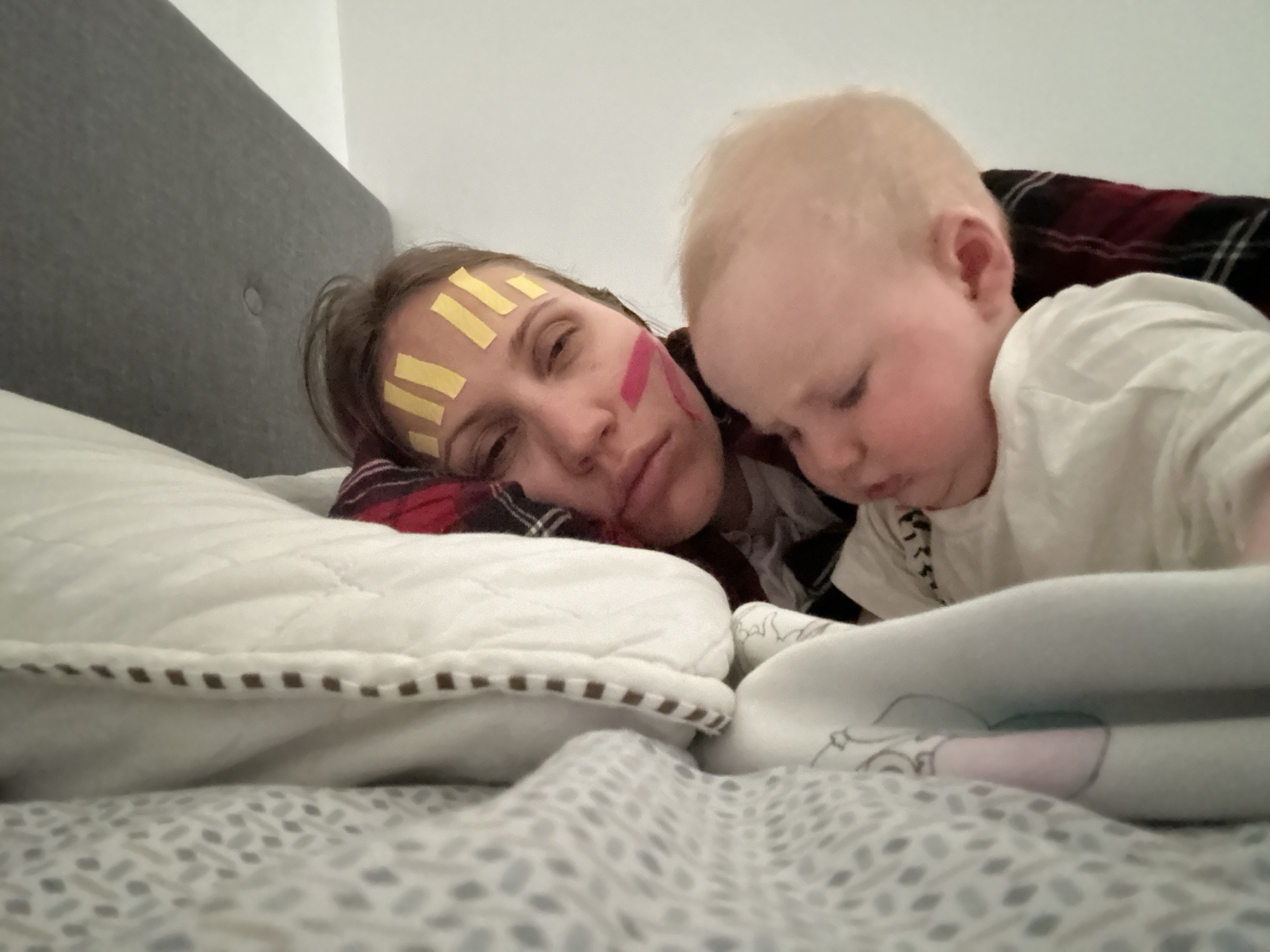 A woman and a baby are on a bed. The woman is lying on a white pillow with brown edges, and she has several yellow strips of tape on her forehead and a pink stripe on her cheek. She appears to be tired or resting. The baby, with blond hair, is positioned next to her, looking downwards. The bedding has a white and grey patterned cover. A red and black plaid blanket is partially visible. The location is Fethiye, Turkey.