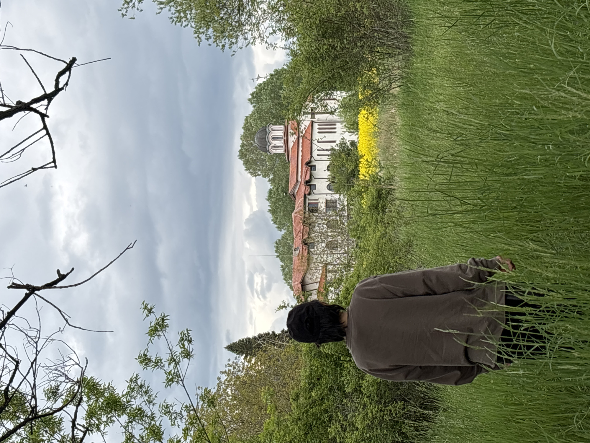 The image depicts a person standing in a field of tall green grass, facing a white building with a red roof and a black dome, partially obscured by trees. Yellow flowers are visible behind the building. The sky is cloudy. Branches from trees are in the foreground on the left side of the image. The person is wearing a brown shirt or jacket. The location is likely near the outskirts of Sofia, Bulgaria, based on the architecture of the building and the surrounding landscape.