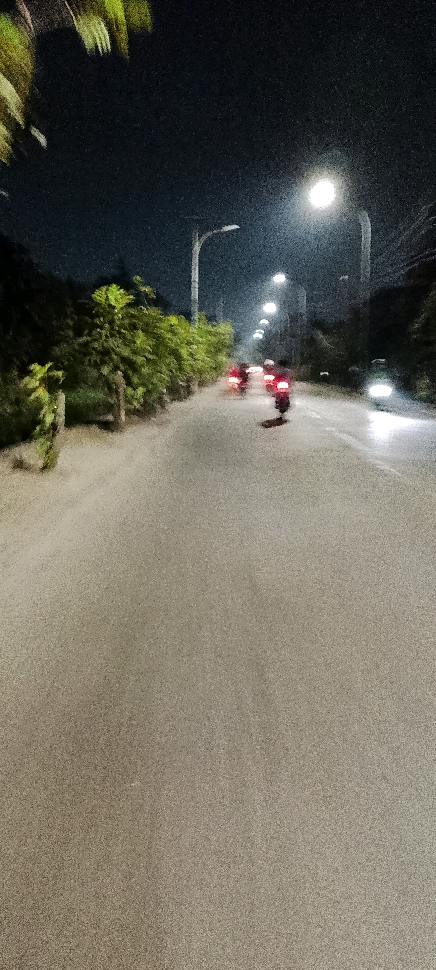 The image depicts a road at night, likely in Bekasi, Indonesia, illuminated by streetlights. Several motorcycles are visible on the road, with their taillights creating streaks of red due to motion blur. The road surface appears to be asphalt or a similar material. Dense foliage lines the left side of the road, with wooden posts interspersed along the edge. Tall, modern streetlights with multiple lamp fixtures are spaced along the right side of the road, casting pools of light and contributing to the overall nighttime ambiance. The sky is dark, indicating it is nighttime. The image captures a sense of movement and activity on the road.