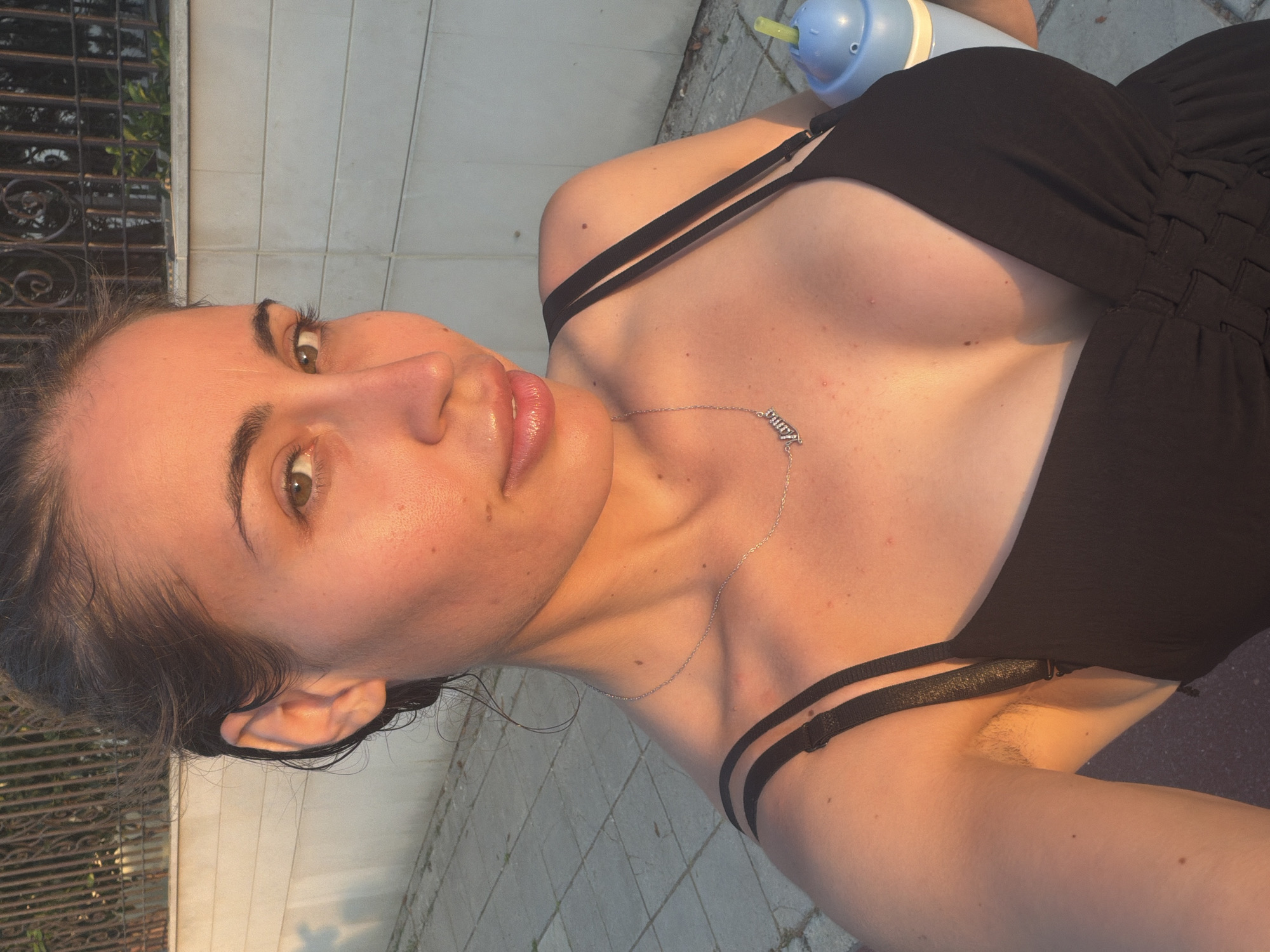 A young woman of Mediterranean descent, with dark hair pulled back and a light complexion, stands outdoors in what appears to be a residential area, possibly Pervolia, Cyprus. She is wearing a black, low-cut dress with thin straps and a silver necklace with a nameplate that reads "Anna." The sun is setting, casting a warm glow on her face and décolletage, highlighting her features and her smooth skin with a few scattered freckles. Her hair is slightly damp, suggesting she may have recently been swimming or exercising. She is holding a blue sippy cup with a straw in her right hand. In the background, a paved walkway is visible, bordered by a light-colored wall and a dark metal fence. The overall impression is one of a casual, relaxed moment captured in the late afternoon light.