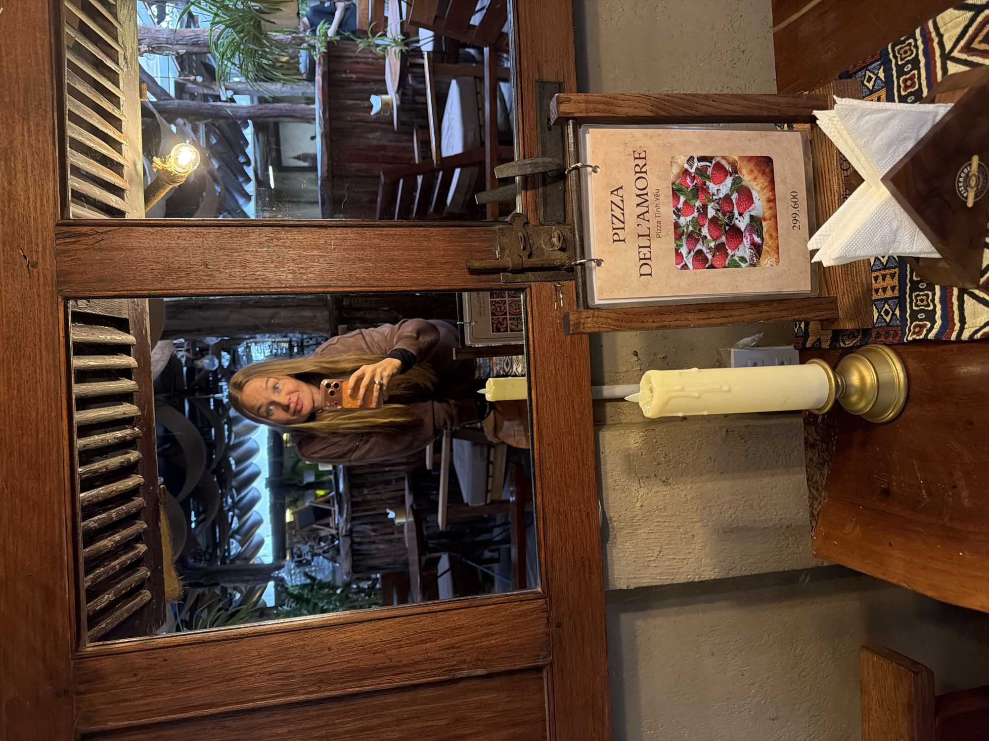 A woman with long blonde hair is taking a selfie in a mirror. She is wearing a brown long-sleeved shirt and holding an orange smartphone. She is in a restaurant with a rustic interior, featuring wooden furniture and decorative lighting. A menu for "PIZZA DELL'AMORE" is displayed on a wooden stand in front of a stack of white napkins. The menu shows a picture of a pizza topped with strawberries. The restaurant appears to be located in Lâm Viên - Đà Lạt, Vietnam, as indicated by the Vietnamese text on the menu. There are other tables and chairs visible in the reflection, suggesting a dining area. Two artificial candles in golden candle holders are also present in the foreground. The overall atmosphere suggests a casual dining experience.