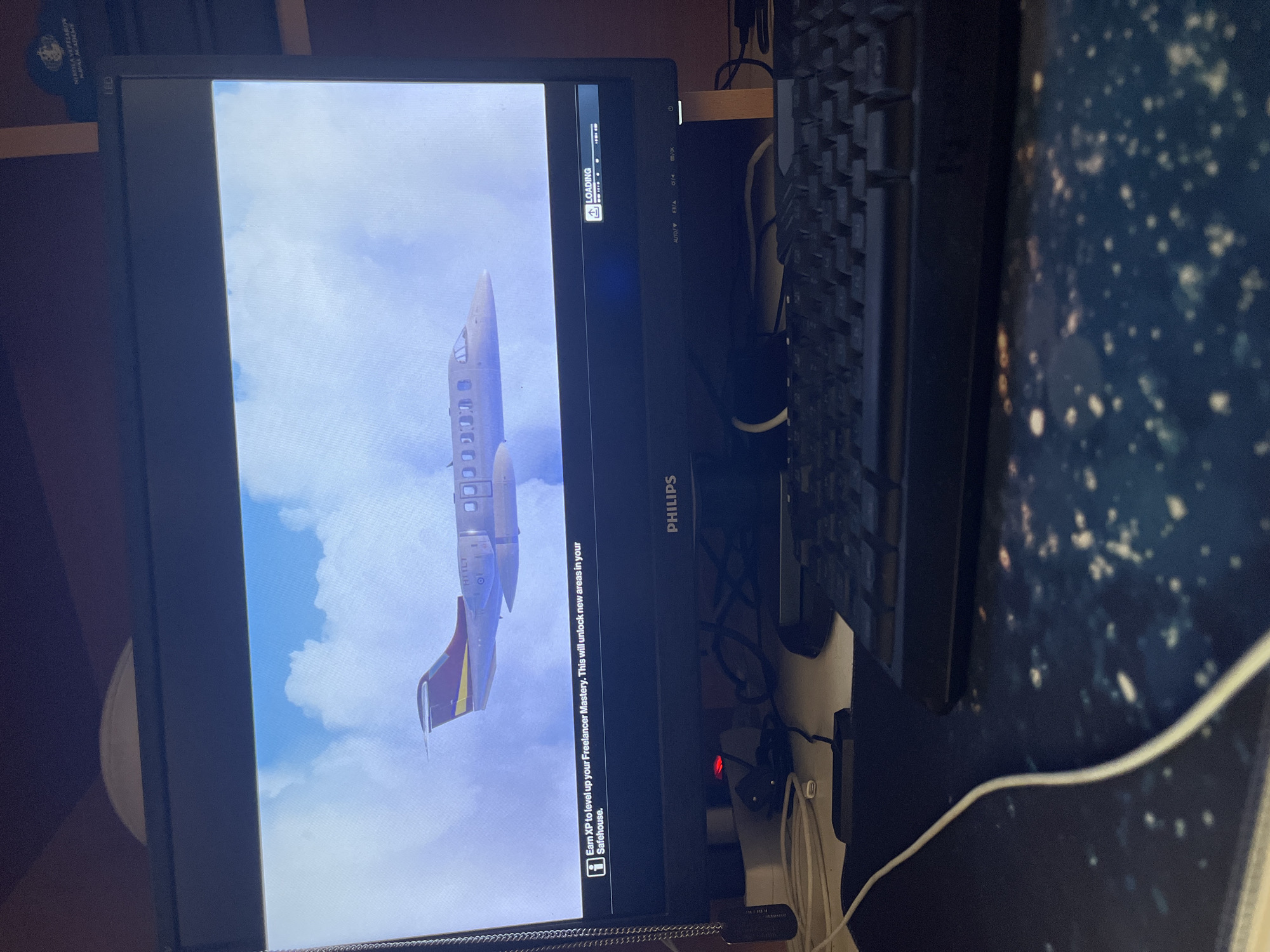 A personal computer setup is displayed indoors, viewed from an angled, slightly elevated perspective. The main subject is a black Philips computer monitor, oriented vertically, displaying a video game loading screen. The screen shows a private jet or small passenger plane flying against a bright blue sky with white clouds. In the upper left corner of the game screen, the word "LOADING" is visible alongside a progress bar and two small icons. At the bottom of the game screen, an informational banner reads: "Earn XP to level up your Freelancer Mastery. This will unlock new areas in your Safehouse." The plane has text resembling "H1TL7" on its tail.
To the right of the monitor, a black keyboard is partially visible on a dark blue/black mouse pad with a speckled, possibly cosmic design. A white cable runs across the mouse pad. Behind the monitor, a cluttered workspace is visible, including various tangled wires, a power strip with a red indicator light, and a white Samsung USB drive plugged in. On a shelf above and to the left of the monitor, an object bears the text "NIKOLA VAPTSAROV NAVAL ACADEMY," indicating a connection to Varna, Bulgaria. The monitor itself has "PHILIPS" printed on its bezel, along with "LED" and various control button labels such as "AUTO/". The scene is well-lit, likely by indoor artificial light.