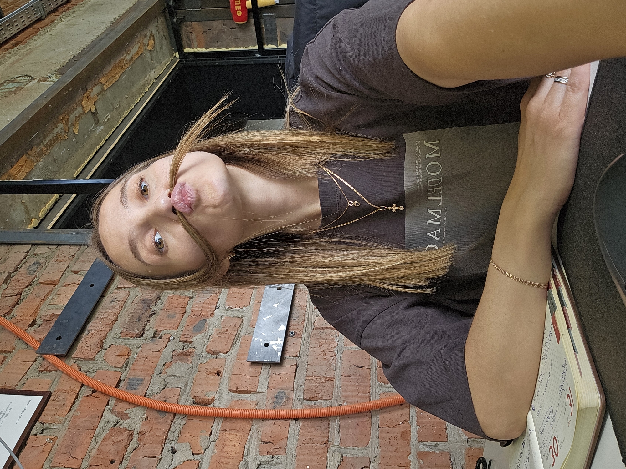 The image depicts a young woman playfully posing indoors in Chaikovsky, Russia. She is shown from the chest up, looking directly at the camera with a lighthearted expression. Her lips are pouted into a kissy face, and she is holding a strand of her long, light brown hair to mimic a mustache. She has brown eyes.
She is wearing a dark gray short-sleeved t-shirt. Visible text on her shirt reads "MODERN" printed vertically, with smaller text "LIVE VIVACIOUSLY" horizontally below it. She accessorizes with a gold-colored layered necklace featuring two pendants: one resembling an infinity symbol or a looped chain, and the other a cross. A simple gold chain bracelet is on her left wrist, and she wears a patterned ring on her right ring finger. Her right arm rests on an open book or journal, displaying large numbers "30" and "31," likely indicating dates, with "янв" (Jan) partially visible next to "31." Some handwritten, illegible notes are also present on the pages.
The background features an exposed red brick wall on the lower half, with an orange corrugated electrical conduit running horizontally across it. A dark metal bracket is mounted on the brick. Above the brick wall, an industrial-style ceiling or upper wall section is visible, characterized by dark beams and exposed spray foam insulation. The setting suggests a casual interior space, possibly a cafe or restaurant with a rustic or industrial aesthetic. The overall mood is playful and informal.
