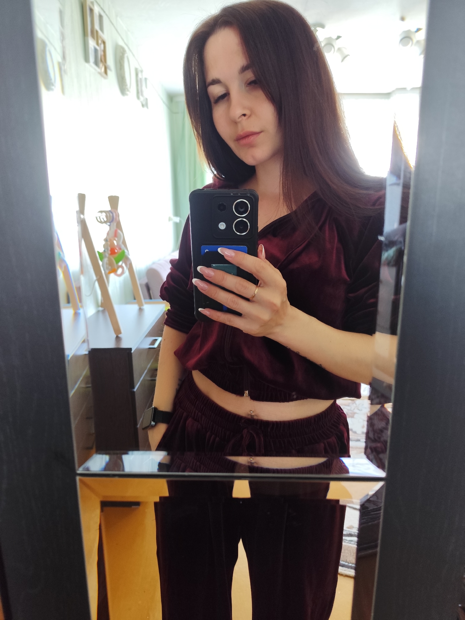 A young woman is taking a selfie in a mirror. She has long, brown hair and is wearing a maroon velvet tracksuit consisting of a cropped jacket and pants. A small section of her stomach is visible. She is holding a black smartphone in her right hand, which is decorated with cards and a ring on her ring finger. The mirror is framed in black. The background includes a dresser with various decorative items and a bright window. The location is in Фоки, Russia.