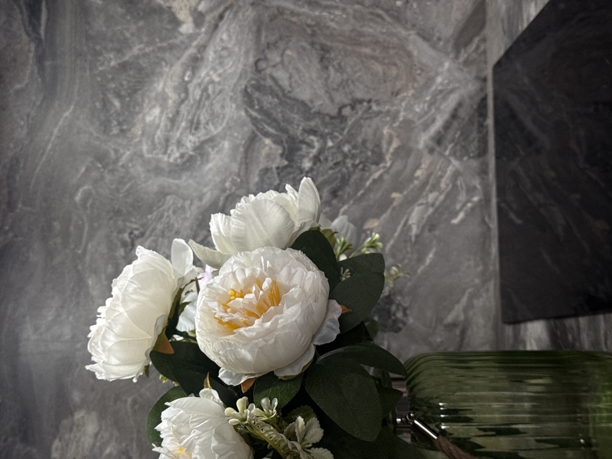 The image displays an indoor still life composition featuring a bouquet of artificial white flowers in a dark green glass vase, set against a marbled grey wall. The main subjects are several large, white, full-bloom flowers, resembling peonies or garden roses, with visible yellow centers and ruffled petals. These are interspersed with dark green leaves and smaller clusters of light green or white filler buds.
The flowers are arranged in a dark green, transparent glass vase that features horizontal ridging, particularly noticeable on its upper portion which is visible in the lower right corner of the frame. To the right of the bouquet, a dark, flat, rectangular object, possibly a panel or shelf, partially extends into the shot, contrasting with the patterned wall.
The background is dominated by a wall with a distinctive grey and white marbled or stone-like texture, exhibiting swirling patterns and lighter veins. The lighting appears soft, creating gentle shadows, particularly on the right side of the image and within the vase, suggesting an interior setting with either natural indirect light or artificial illumination.
There are no people or any specific activity taking place; the scene is purely decorative. The overall impression is one of modern elegance and tranquility. No visible text is present in the image. The location is an interior space, and the time of day or weather conditions cannot be determined.