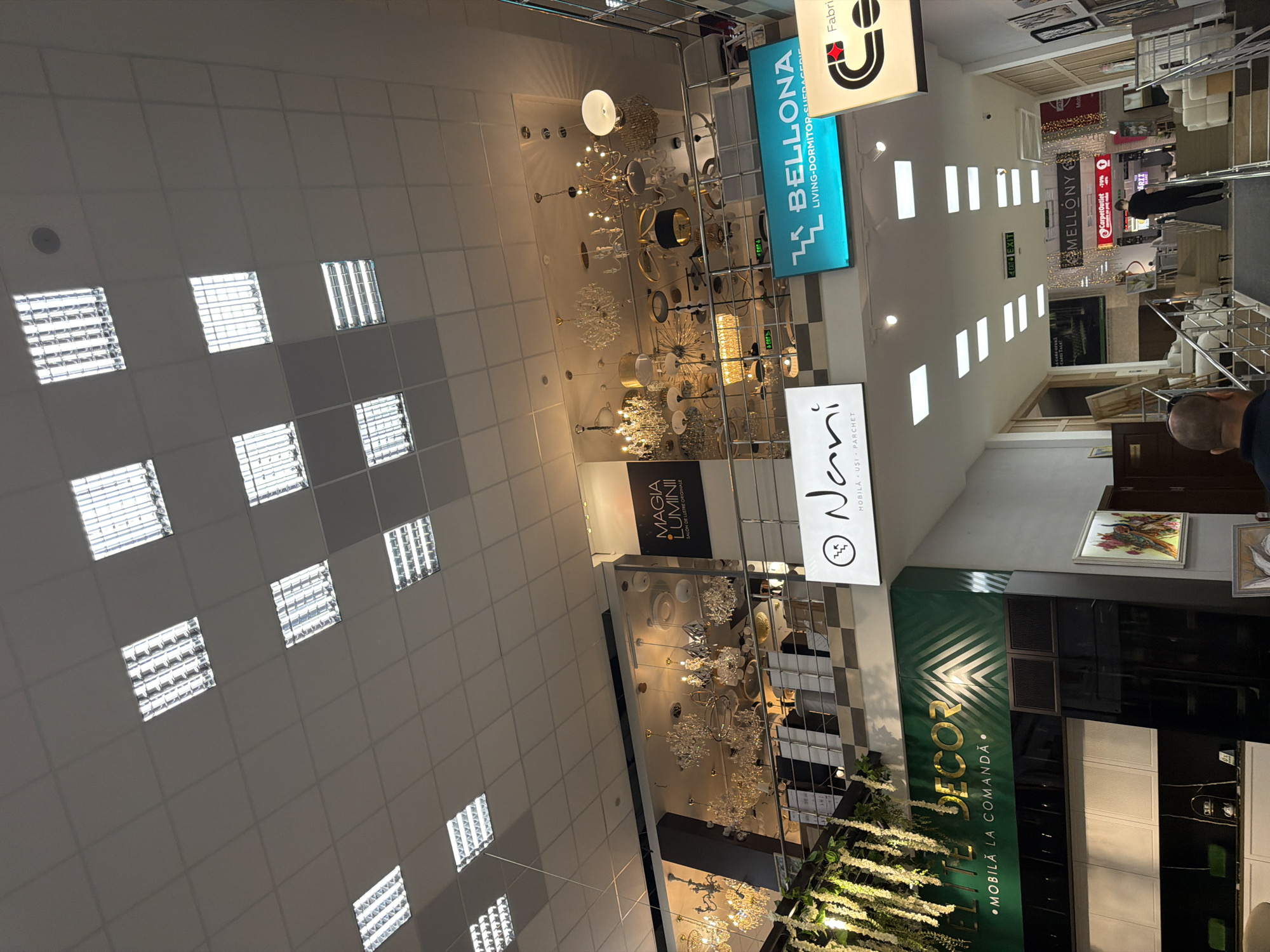 The image depicts the interior of a large retail store, likely a home furnishings or lighting showroom, situated in Trușeni, Moldova. The ceiling is adorned with numerous fluorescent light fixtures arranged in a grid pattern. Below the ceiling, a mezzanine level displays an array of ornate chandeliers and other decorative lighting fixtures. Various signage is visible throughout the store, including "MAGIA LUMII," "BELLONA LIVING-DORMITOR-SUFEDERIE," "Nani," "ELITE DECOR," and "MELLONY." A few individuals are present in the store, including a person wearing a black t-shirt and mask standing on a staircase in the background. The environment appears to be well-lit and organized, typical of a retail establishment.