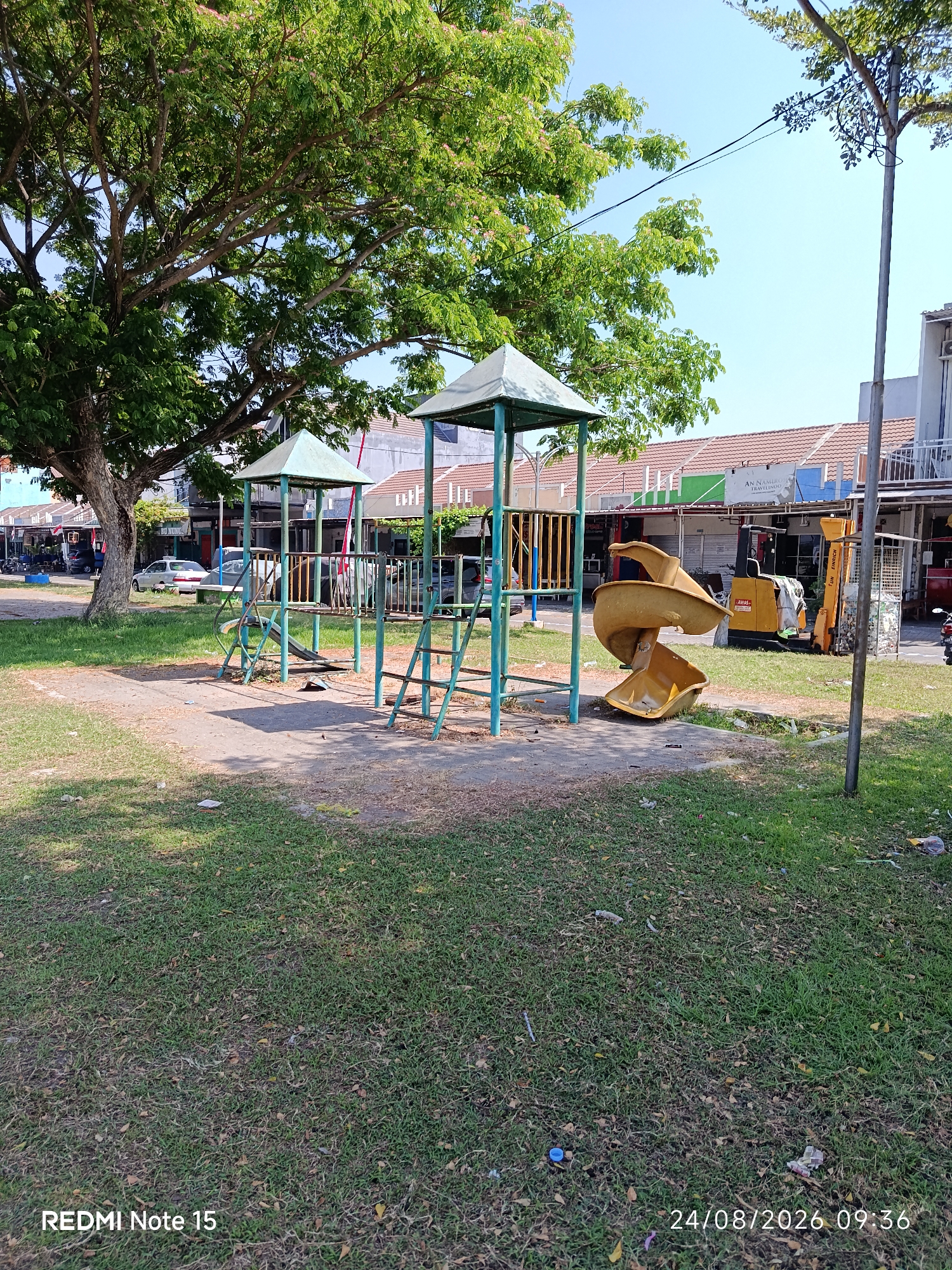 The image depicts a public playground area with a large tree on the left and a row of shops in the background. The playground features two small structures with green metal frames and conical roofs, one with a slide. A yellow spiral slide is also present in the foreground. The ground is a mix of pavement and patchy grass. In the background, a forklift is parked in front of the shops. The scene appears to be located in the city of Keboananom, Indonesia, as indicated by the timestamp "24/08/2026 09:36" and the text "REDMI Note 15" which might imply the device used to capture the image, potentially associated with local context. There are no people visible in the image. The environment seems to be a somewhat neglected public space, with scattered debris on the grass.