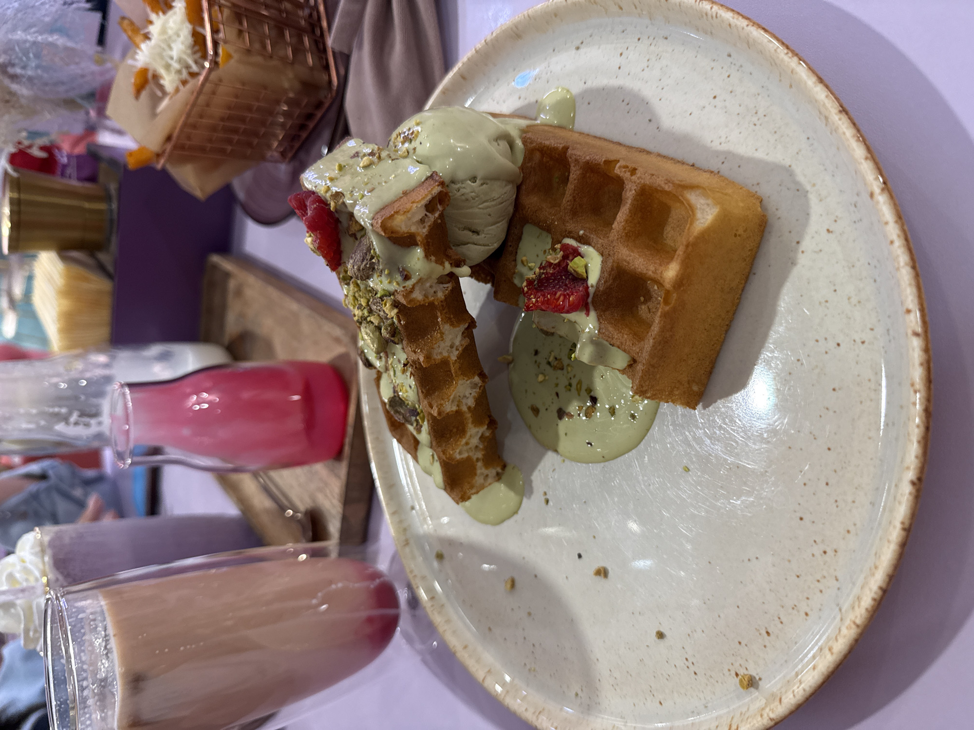 The image depicts a waffle dish with green ice cream and pistachio toppings on a speckled white plate. A small basket of fries with shredded cheese is visible in the background, along with glasses of various beverages, including a pink drink and a brown drink. The scene appears to be indoors, possibly in a cafe or restaurant. Information about Vladivostok, Russia is not discernible from the image.