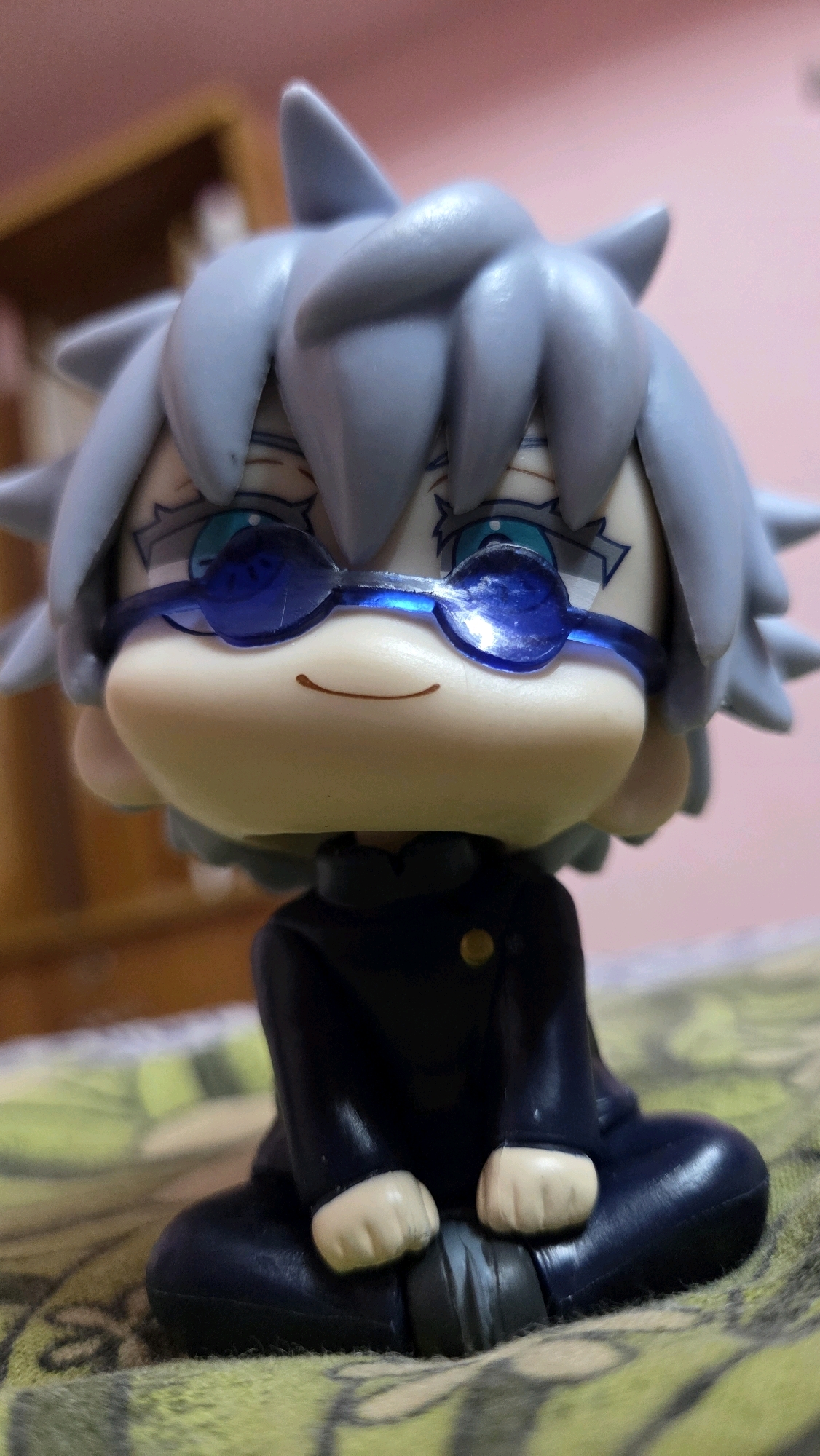 A close-up, eye-level shot features a stylized anime figurine of a character with spiky light grey hair and blue-tinted glasses. The figurine has a light-skinned face with a small, subtle smile and detailed blue eyes. It is wearing a dark blue uniform with a golden button. The figurine is posed in a seated position, with its hands resting on its lap. The background is softly blurred, showing a pink wall and a wooden shelf. The figurine rests on a patterned surface, possibly a cloth or blanket, with a predominantly green and yellow design. No specific location in Chalakudy, India is identifiable in this image.