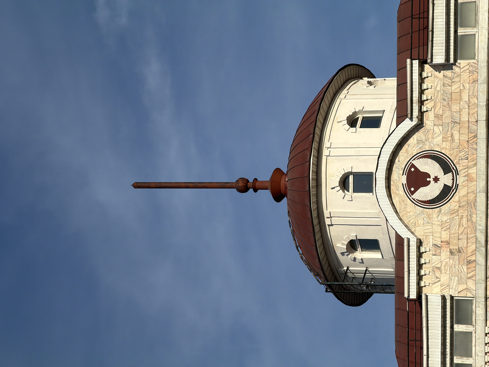 A low-angle, vertically oriented photograph depicts the upper portion of a building against a backdrop of a blue sky with wispy clouds. The architectural style appears to be Soviet-era, possibly representing Stalinist architecture. The building features a dome-shaped top with a reddish-brown roof and a long, pointed spire extending from its center. Three arched windows are visible on the rounded facade beneath the dome. To the right, a portion of the building has a decorative emblem with what seems to be an eagle-like symbol. The building's facade is tiled with a mix of light-colored stone tiles. This building is located in Izhevsk, Russia.