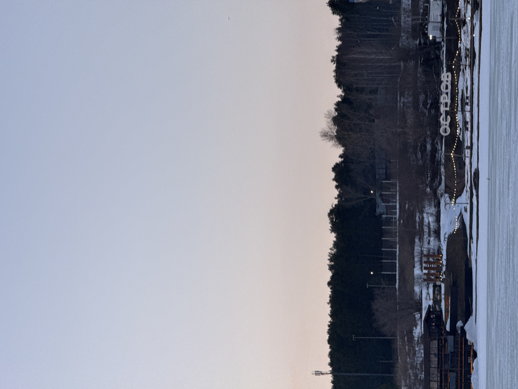 The image shows a view of Votkinsk, Russia, during what appears to be late dusk or early twilight. The left half of the image is predominantly a pale sky, suggesting the sun has set or is very close to setting. On the right side, a shoreline is visible, possibly part of a lake or reservoir, with a frozen surface covered in snow. A line of trees borders the shoreline, becoming denser towards the background. There are structures along the shoreline, including buildings, possibly part of a recreational area or park. Illuminated signage in Russian is present, along with decorative lighting. The word written in Russian is "Остров", which means "Island" in English. The scene is quiet and somewhat bleak due to the low light and winter conditions.