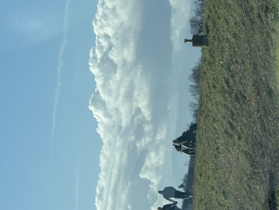 The image displays an outdoor scene in Altenheim, Germany, under a bright blue sky with extensive white cumulus cloud formations occupying the mid-section. A faint contrail is visible in the upper blue sky. The foreground and midground consist of a green, grassy slope with patches of brown. On the left, two individuals are walking from left to right, with their backs partially visible. The person on the left wears dark clothing and carries a shoulder bag. The person on the right wears a light-colored jacket and dark pants; their hands appear joined. Centrally, two individuals are seated on a dark wooden bench with large, circular side supports, facing away from the viewer and positioned on the crest of the grassy area. To the right, a weathered, cylindrical stone or concrete pillar, topped with a metal T-shaped fixture, is embedded in the grassy slope. A distant line of leafless trees and a hazy, undulating landscape form the background beneath the clouds.