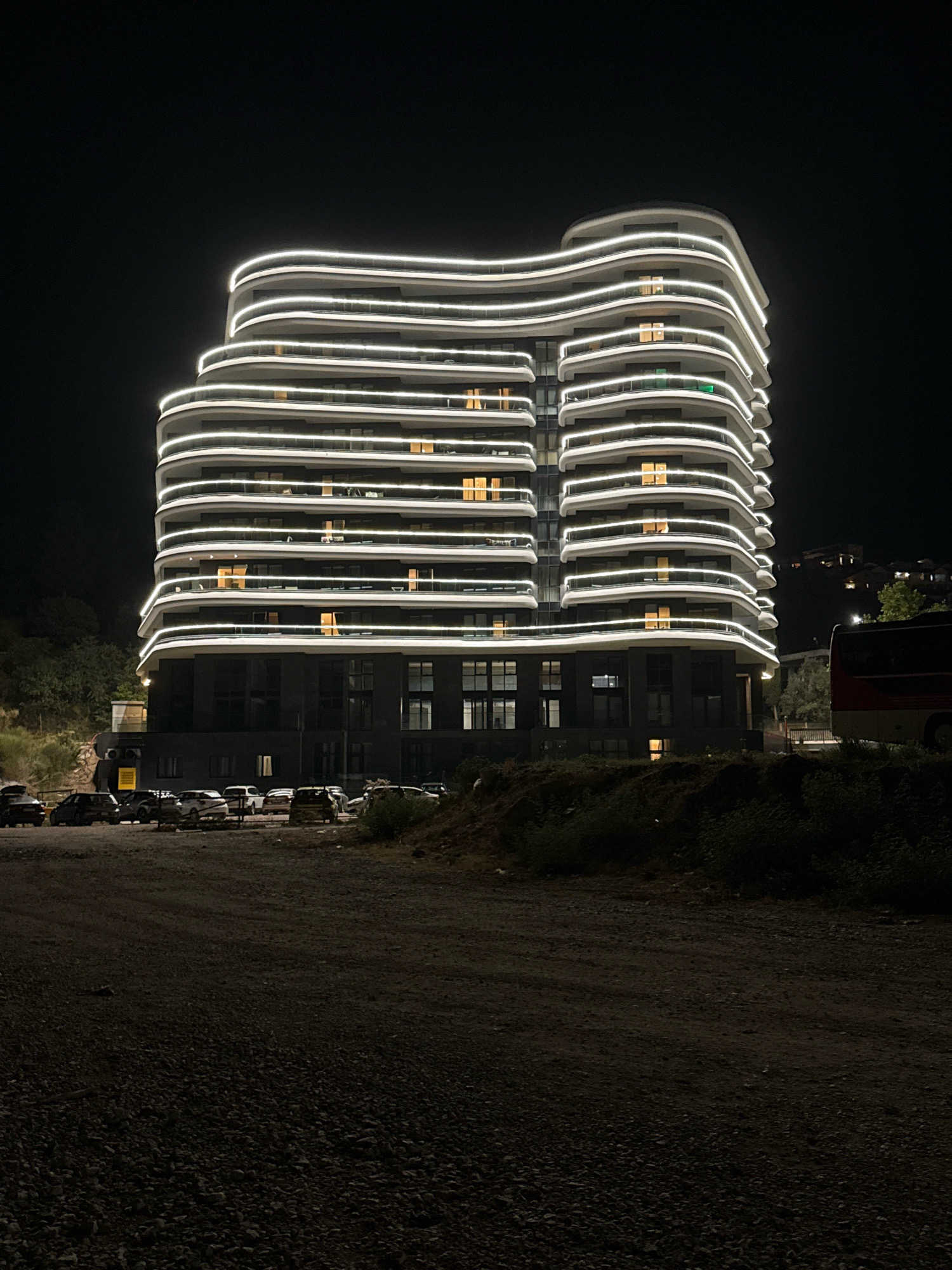 An exterior, eye-level, nighttime photograph captures a modern, multi-story building with a unique, wavy facade illuminated by continuous white LED strip lights along its curved edges. The building is situated in Boreti, Montenegro, appearing to be a residential or hotel complex. Below the building, a gravelly, unpaved lot is visible, with several parked cars arranged in a row. A red and white bus is partially visible to the right of the frame, and the surrounding landscape is dark with trees and a hill visible in the background. The overall impression is one of contemporary architecture against a natural, dimly lit setting.