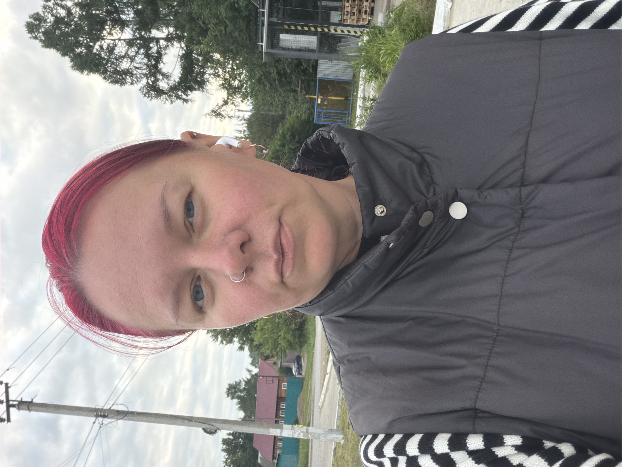 A woman with bright pink hair stands outside, facing the camera. She is wearing a dark gray puffy vest over a black and white striped long-sleeved shirt. She has a silver hoop nose ring and a silver earring in her right ear, along with a white earbud in her left ear. Her left cheek shows a slight blush.
The background suggests a semi-rural or suburban environment. To the left, a utility pole with wires is visible, along with a green fence and houses with red roofs. To the right, there are trees, some green foliage, and a bus stop shelter with a metal frame and some wooden pallets stacked nearby. The sky is overcast with gray clouds. The overall setting appears to be the city of Chaikovsky, Russia, based on the presence of residential buildings and infrastructure typical of a Russian town.