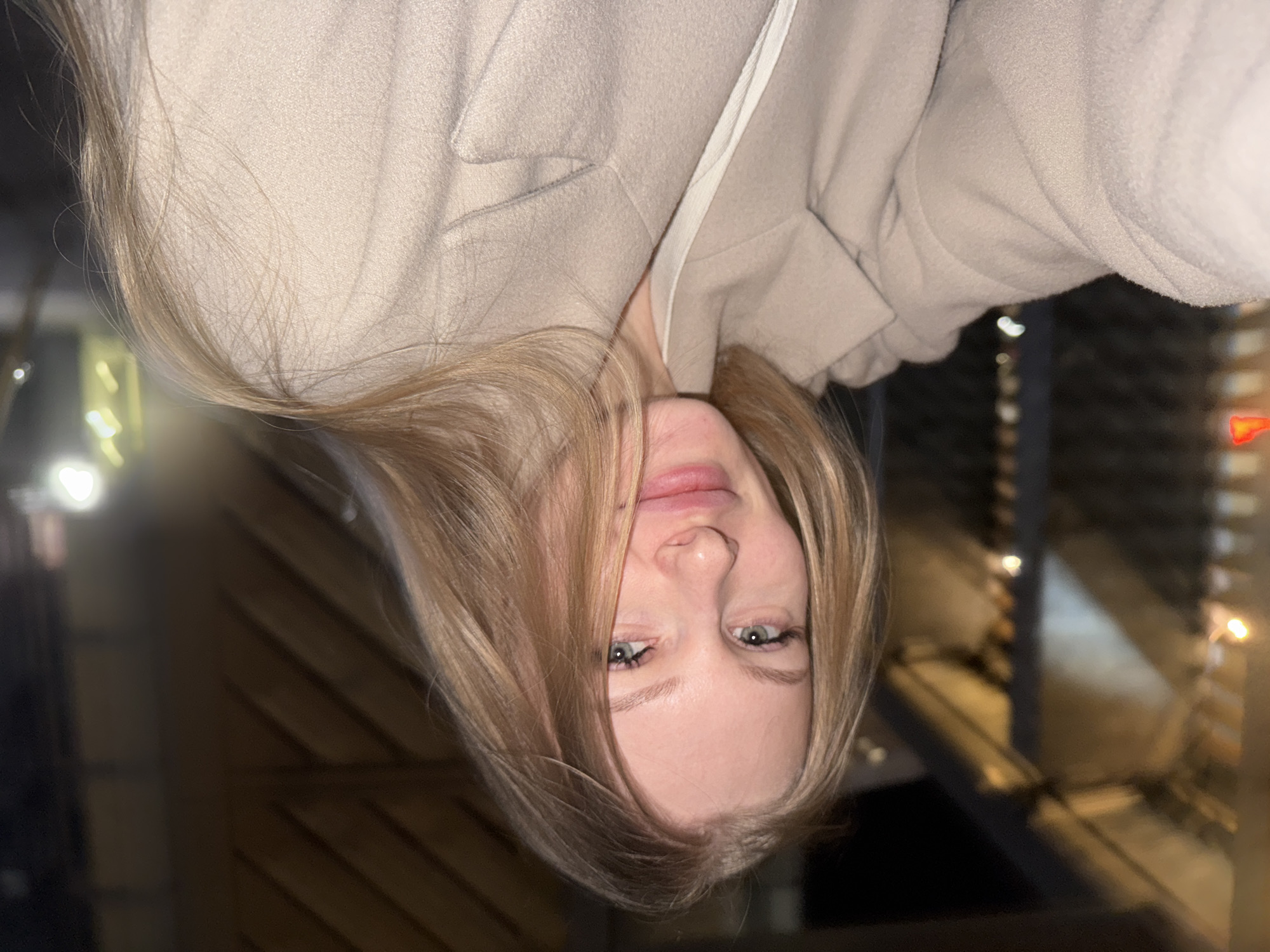 This media file presents a close-up portrait of a young woman, captured against a blurred, dark background. The subject has long, straight to slightly wavy blonde hair that frames her face, fair skin, and bright blue-green eyes with a direct gaze towards the camera. Her expression is calm and neutral, with her lips slightly parted. She is wearing a warm, light beige or cream-colored coat, possibly made of wool or a similar textured material, with a white collar or undershirt visible near her neck.
The background is dark and out of focus, featuring numerous glowing bokeh lights in shades of white, yellow, and orange. This suggests an urban or indoor setting at night, with hints of structural elements, possibly railings or windows, faintly visible in the soft blur. The lighting on her face appears to be artificial, illuminating her features against the dim backdrop. The photograph is presented with a vertical flip and 90-degree rotation, showing the woman's head upside down, but her gaze remains direct, implying a close-up portrait or selfie. The setting for this image is Perm, Russia.