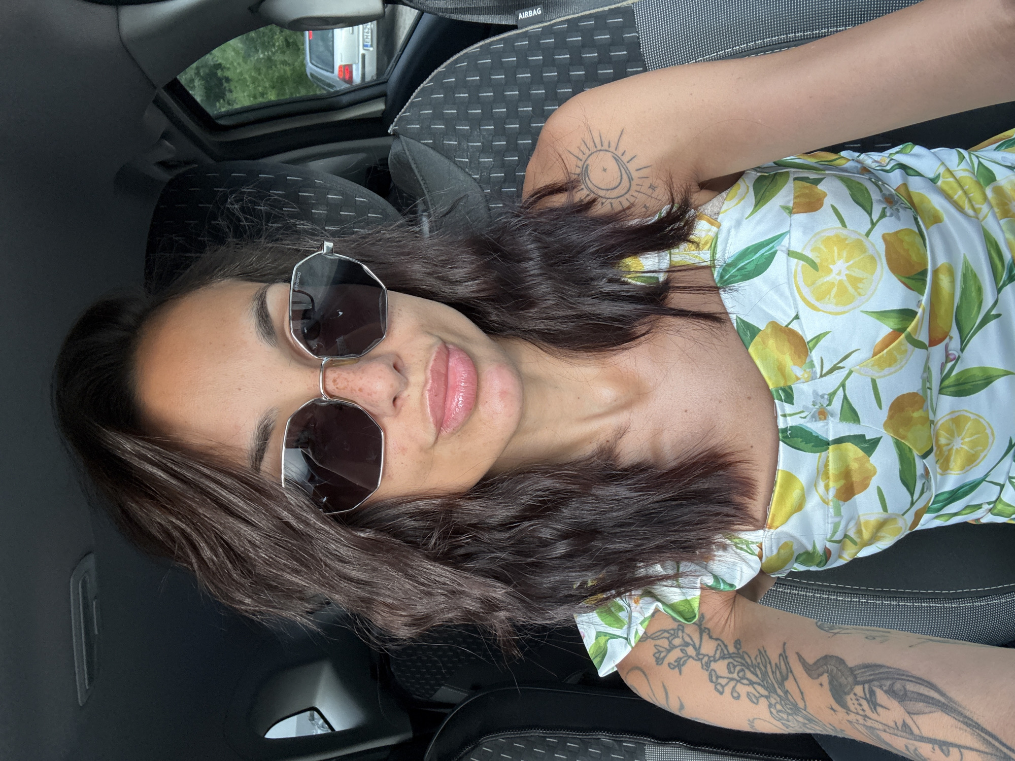 A woman is sitting in the driver's seat of a car, taking a selfie. She has medium-length, dark brown, wavy hair and is wearing large, hexagonal, silver-rimmed sunglasses with dark lenses. She has freckles across her nose and cheeks and is wearing a strapless dress with a white background and a pattern of yellow lemons and green leaves. She has tattoos on her left arm and right shoulder. The car's interior is visible, with dark fabric seats and the dashboard. Outside the car window, a silver car is visible on a tree-lined road, suggesting the car is in a suburban or rural area. The image was taken on a bright, sunny day. The location is the city of Pleven, Bulgaria.
