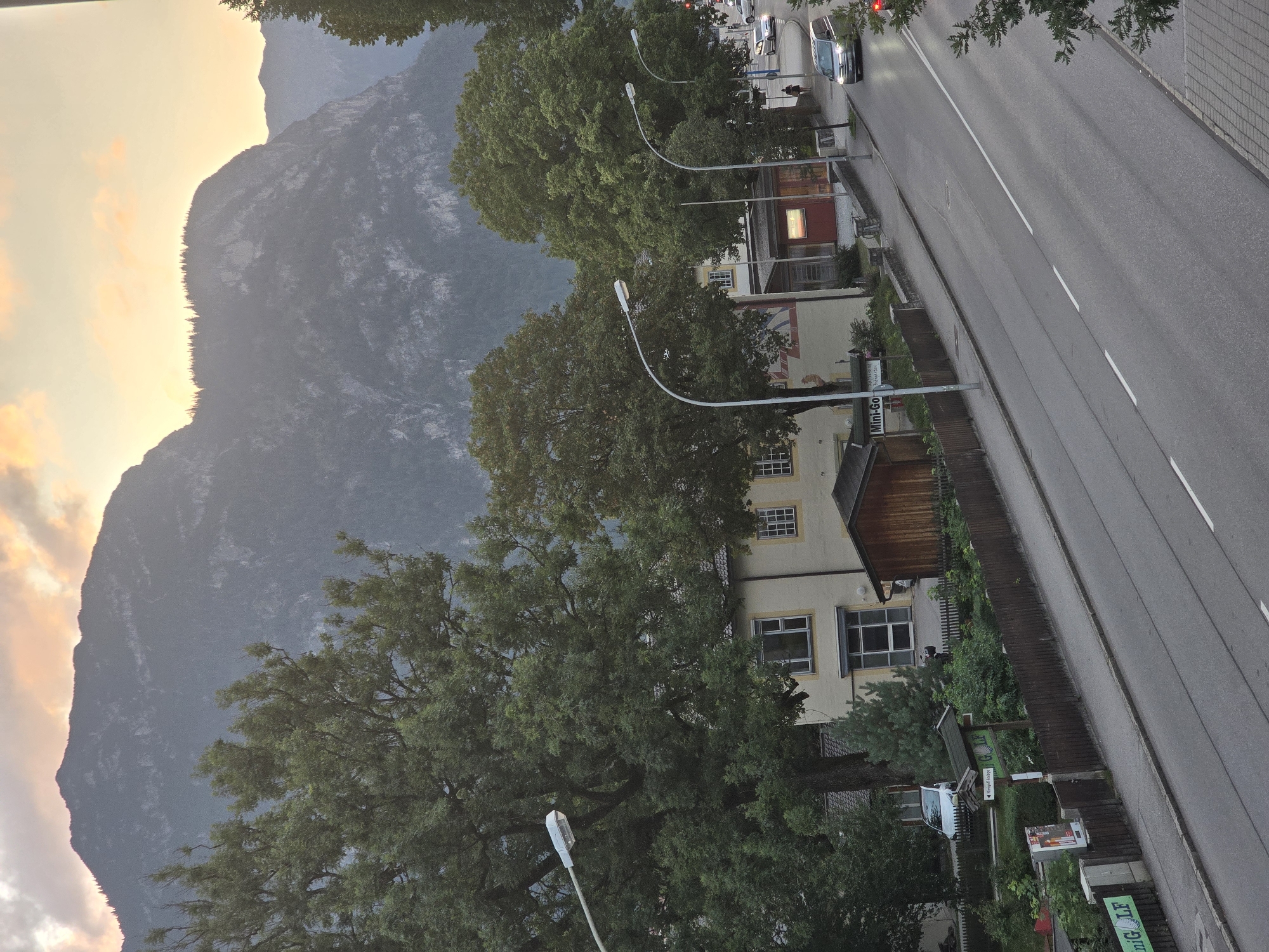 The image shows a street scene in Garmisch-Partenkirchen, Germany, with a mountain range in the background. The sky is partly cloudy with hues of orange and yellow, suggesting it is either sunrise or sunset.
In the foreground, a street runs diagonally across the frame, with cars driving on it. On the left side of the street, there is a sidewalk and a fence enclosing a property with trees and a building. A sign reads "Mini-Golf". On the right side of the street, there are more trees and buildings, including a small red structure with illuminated windows.
Further back, a large, multi-story building with a light-colored facade is visible, partially obscured by large trees. The mountain range in the background is covered in trees and appears rocky in places. The overall atmosphere is calm and serene.