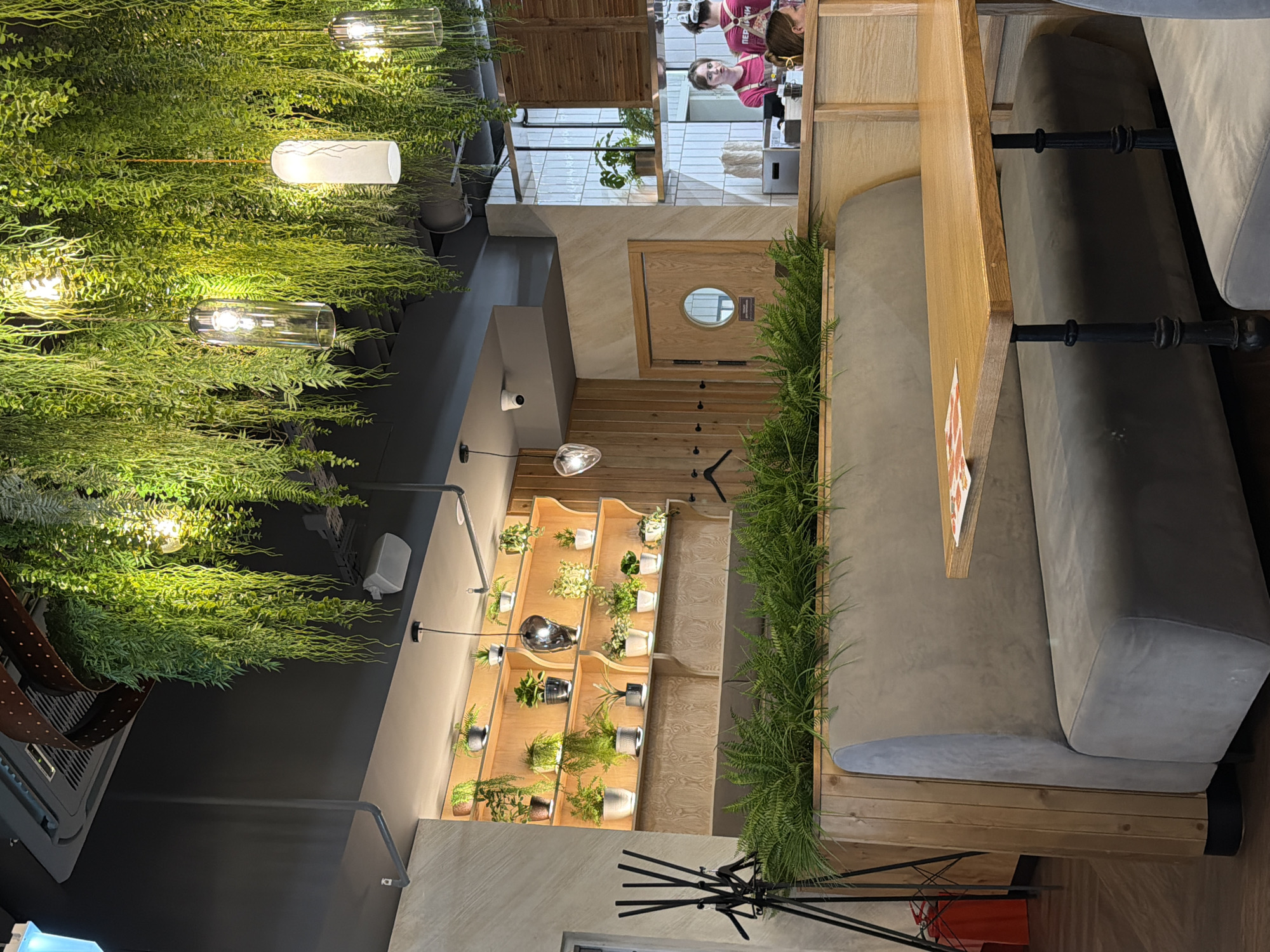 The image depicts an indoor scene, likely a cafe or restaurant, with a strong emphasis on greenery. Hanging plants cascade from the ceiling, interspersed with modern pendant lights. In the background, two women are visible behind a counter, appearing to be staff members. One woman, wearing a pink shirt with Cyrillic text "ПЕРЕКРАСНО" (meaning "beautiful" or "wonderfully"), is looking towards the camera and smiling. The other woman, wearing a red shirt, is also looking in the general direction of the camera. The interior features wooden accents, including slatted walls and shelving units filled with small potted plants. A circular porthole door is also visible. The overall atmosphere appears bright and welcoming, suggesting daytime. The location context is Perm, Russia.
