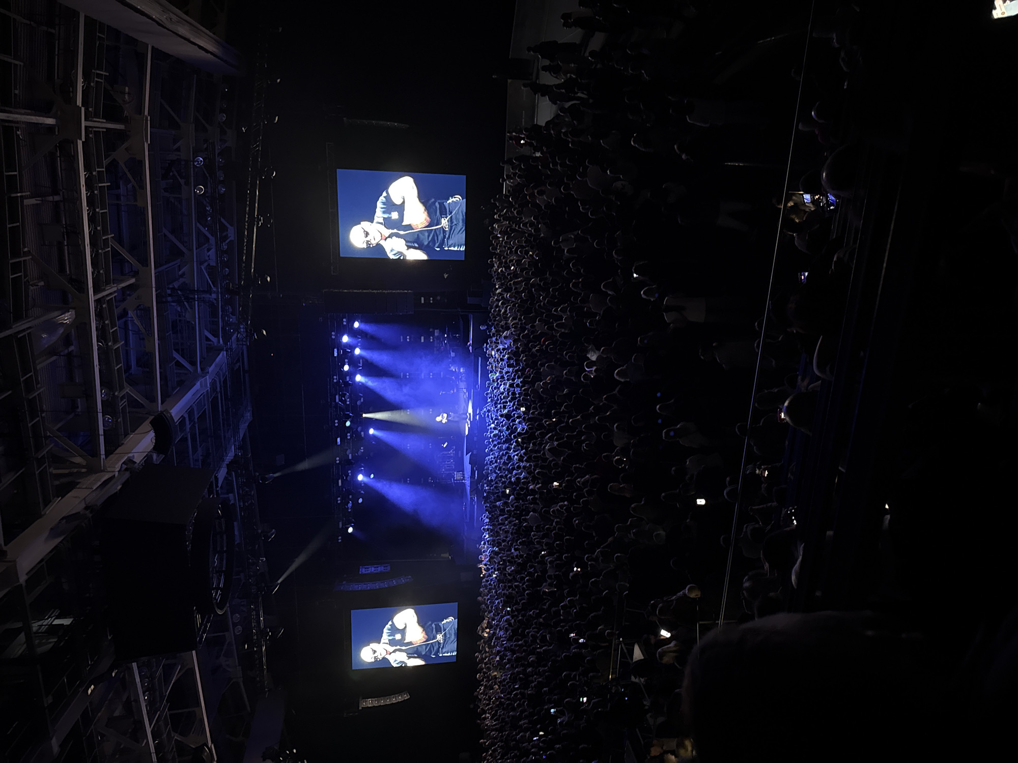 The image shows a concert in Krasnoyarsk, Russia. A large crowd of people are tightly packed together, facing a stage that is lit with bright blue lights. Two large screens flank the stage, showing a performer. Metal scaffolding surrounds the stage and crowd. The right side of the image is very dark, with the crowd densely packed.