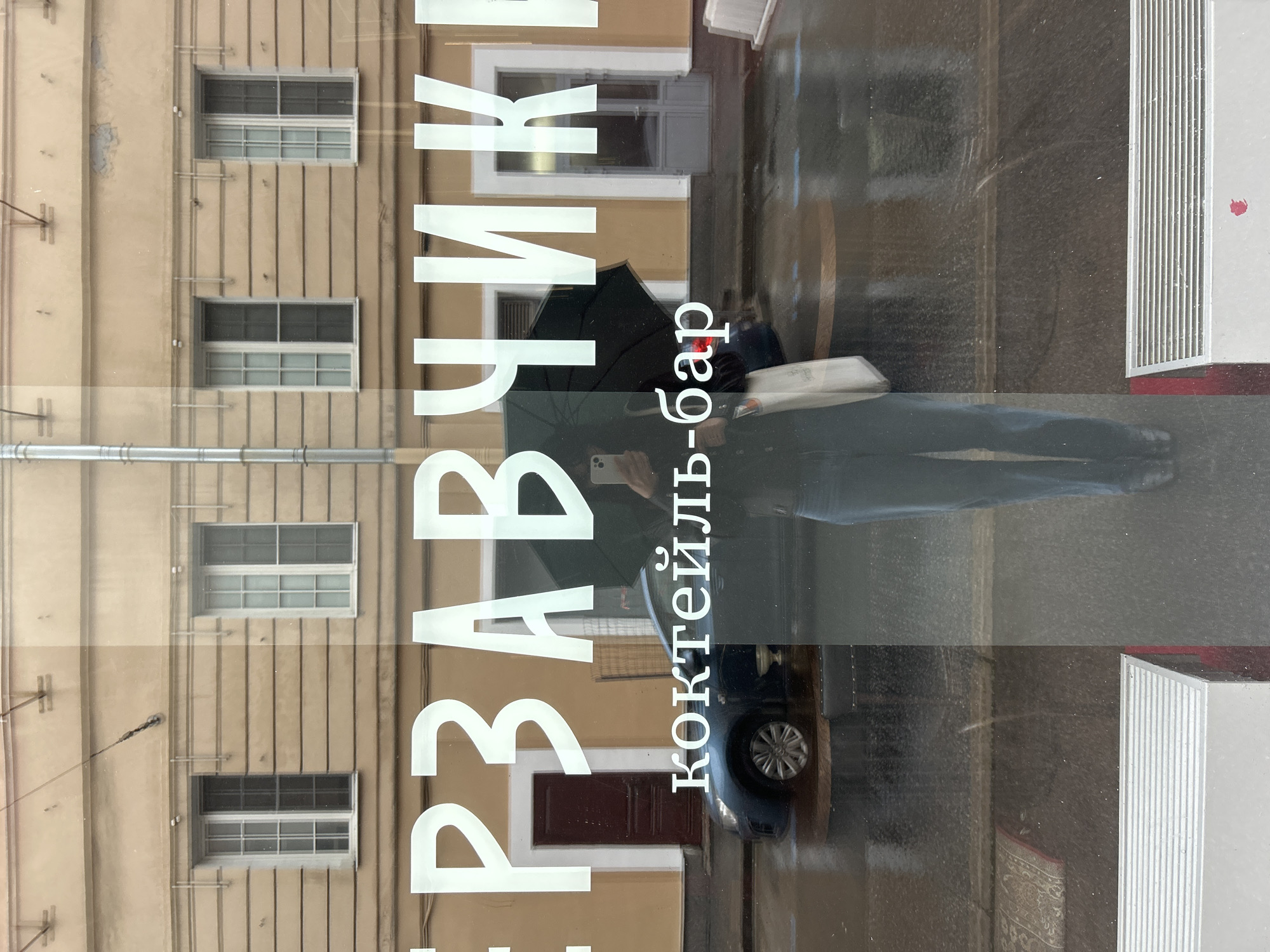 This image depicts a reflection in a window on a rainy day in Saint Petersburg, Russia. The reflection shows a person holding a black umbrella and a white bag, wearing jeans and dark clothing. Behind the person, a blue car is visible on a wet street. The window displays large white Cyrillic lettering that reads "ПЕРЗВУЧИКИ" and "коктейль-бар". In the background, the facade of a building with multiple windows and a brown door is reflected. The overall scene suggests a street-level view of a bar or establishment on a dreary day in the city.
