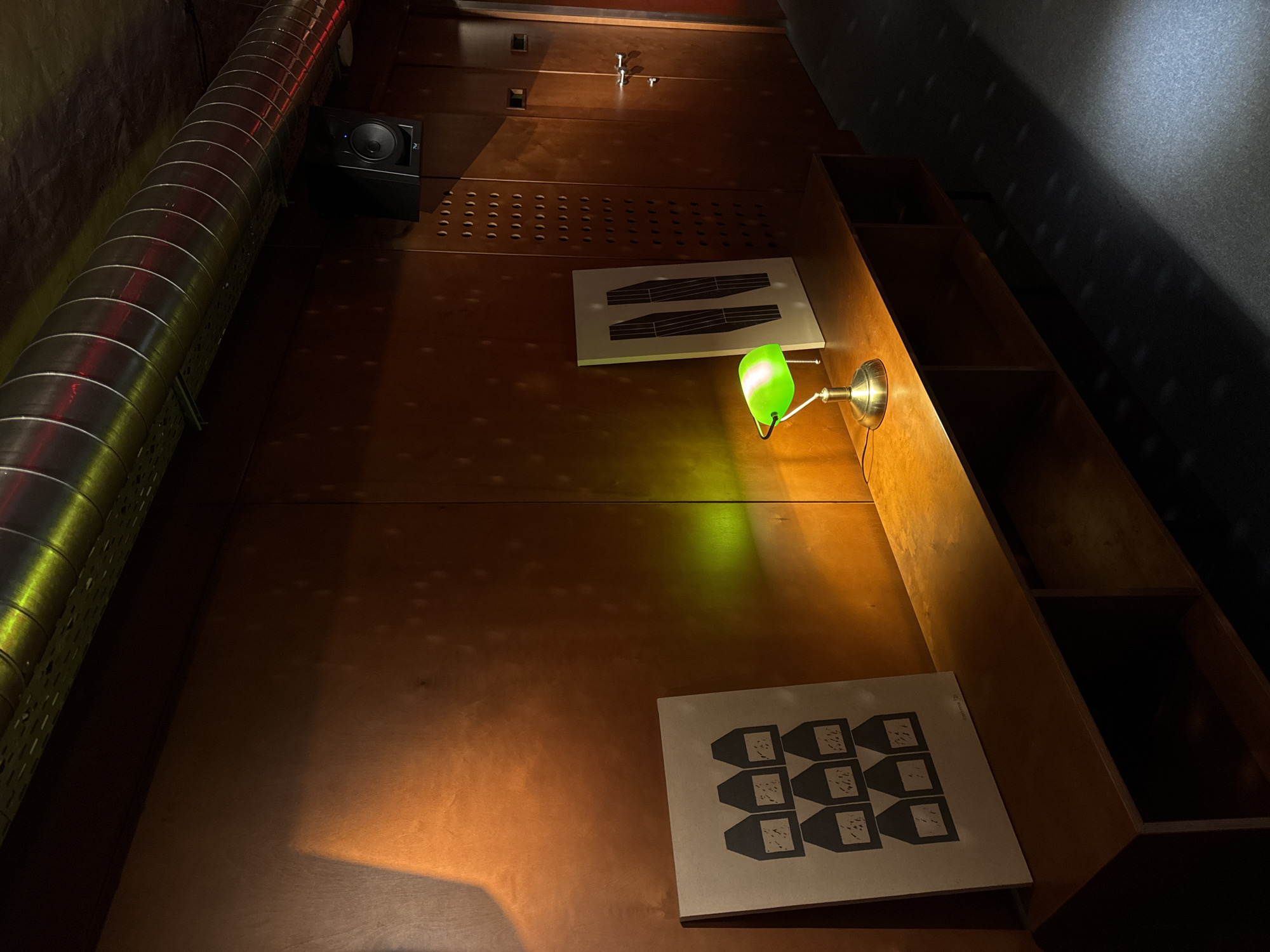 Captured from a low angle, the image portrays a dimly lit interior, likely in Yerevan, Armenia. A prominent metallic duct, exhibiting a corrugated texture, stretches along the upper-left quadrant of the frame. Adjacent to it, a black box, possibly an audio speaker, is affixed to the wall. The wall itself is paneled in a deep, saturated brown wood, featuring rows of ventilation holes and several silver hardware fixtures.
A green-shaded desk lamp casts a concentrated beam of light onto the wall, illuminating two distinct rectangular graphic artworks. One artwork displays a pattern of small, vessel-like shapes arranged in a grid, while the other features a pair of horizontal lines.
To the right, a series of recessed shelves are visible, receding into deep shadow. The lighting in the scene is predominantly dim, with the exception of the concentrated beam from the desk lamp. The ceiling, only partially visible, transitions from dimly lit to entirely obscured in shadow towards the upper-right corner. The composition is characterized by its strong diagonal lines and interplay of light and shadow.