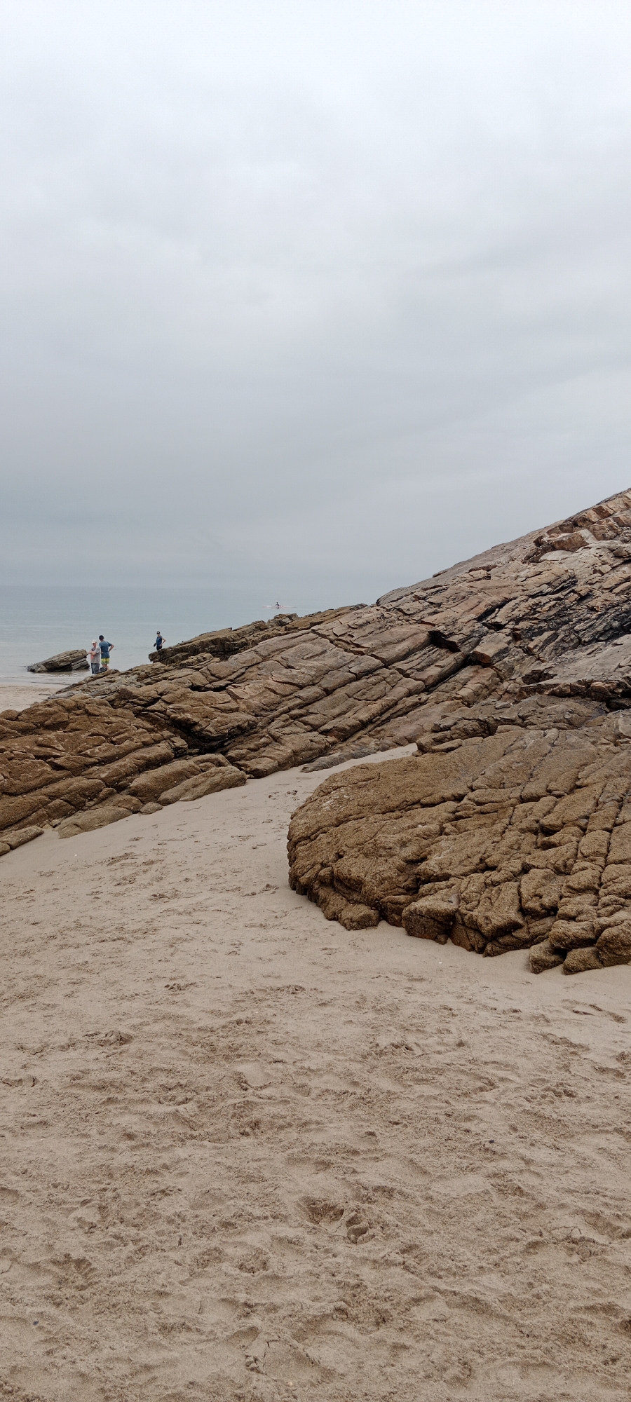 The image shows a sandy beach with large, layered rock formations under an overcast sky. In the distance, a calm body of water meets the horizon. Three individuals are visible standing on the rocks near the water's edge. One person is wearing a blue shirt and shorts, another is wearing a blue shirt and dark shorts, and a third is wearing a dark shirt and shorts. A small boat or kayak is also visible on the water. The sand in the foreground shows footprints.