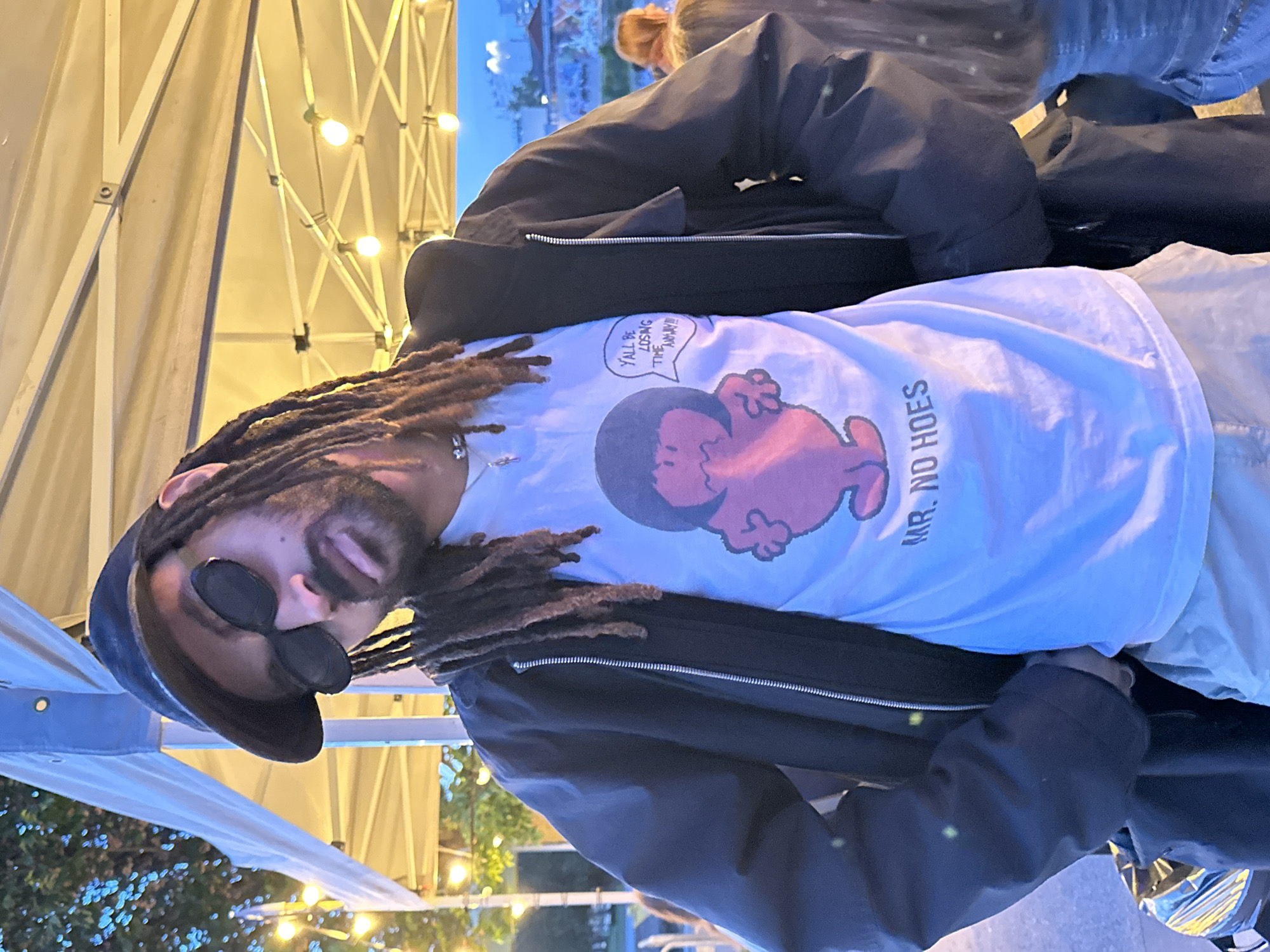 A person with dreadlocks, a goatee, and sunglasses is wearing a hat, a jacket, and a t-shirt with "Mr. No Hoes" printed on it. The person is standing in front of a tent structure with lights, possibly in Paris, France, based on the presence of a building with architecture common to Paris in the background.