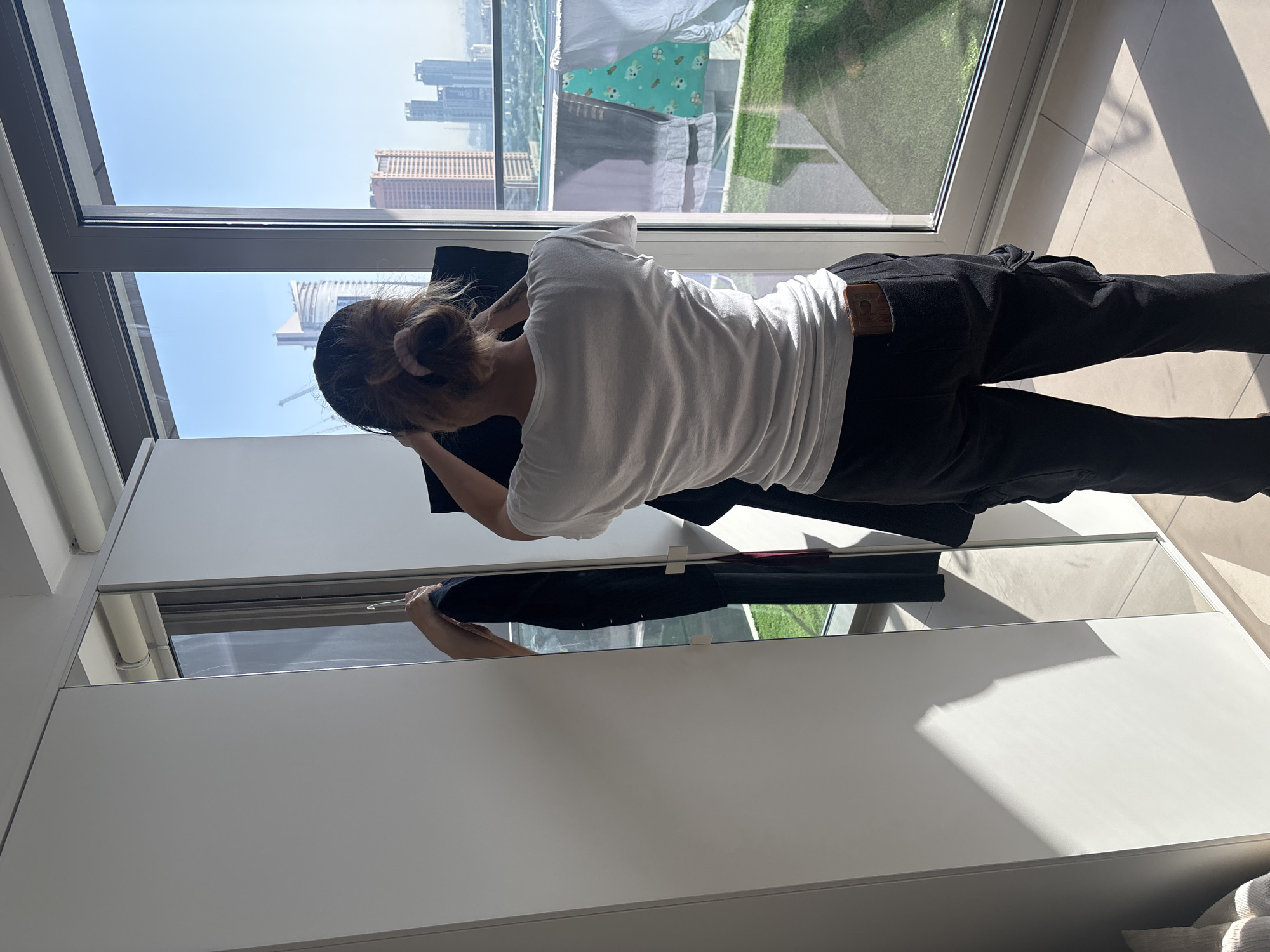 A person stands facing a closet with mirrored doors, holding a dark garment. They are wearing a white t-shirt and black cargo pants, with a mobile phone in their right back pocket. The closet is white and has a sliding door, with a reflection showing more of the closet interior and a glimpse of the person from the side. Sunlight casts a shadow on the closet door. Behind the person, a large window offers a view of a city skyline with tall buildings and cranes, characteristic of Dubai, United Arab Emirates. Clothes hang on a drying rack outside the window, suggesting a residential setting with a balcony or exterior space. The floor is tiled.