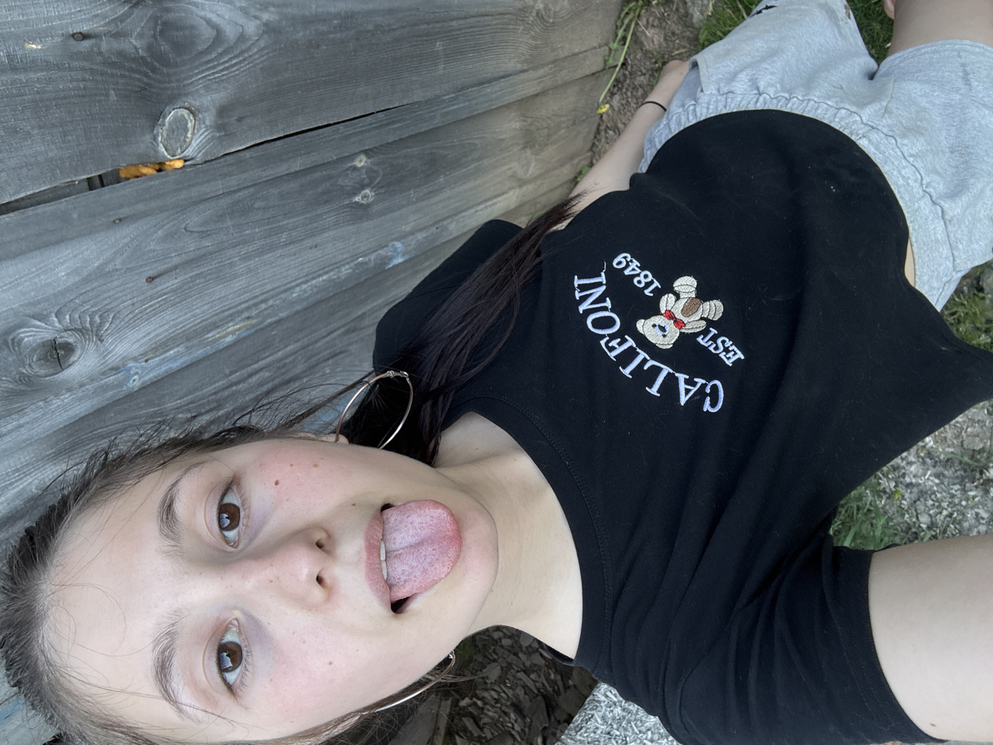A young person is positioned at the bottom of the frame, wearing a black t-shirt with a white embroidered design and grey shorts. The person is lying on their back, near a wooden fence, with a patch of grass and dirt visible in the background. The person is sticking their tongue out, facing the camera. They are wearing large hoop earrings. The location is unspecified in the city of Фоки, Russia.
