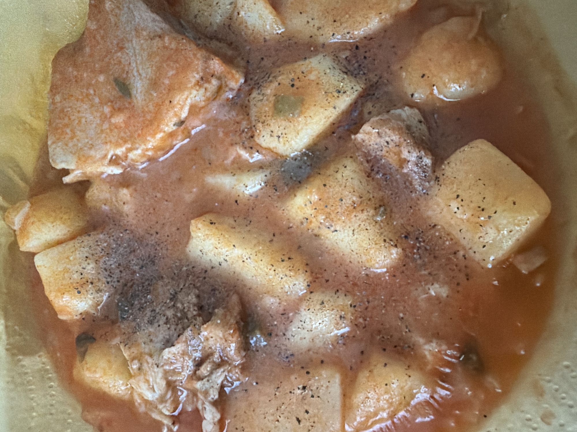 A close-up, overhead shot captures a dish of food, likely a stew or soup, filled with chunks of potato and pieces of meat in a reddish-orange broth. The food is seasoned with black pepper. The dish appears to be served in a light yellow, textured bowl or container, possibly made of paper or a similar disposable material. The background is a solid, light yellow color. There are no people or significant actions depicted. The image is focused on the food itself, with a shallow depth of field. The scene is devoid of any specific geographical markers that would place it in Pazardzhik, Bulgaria.