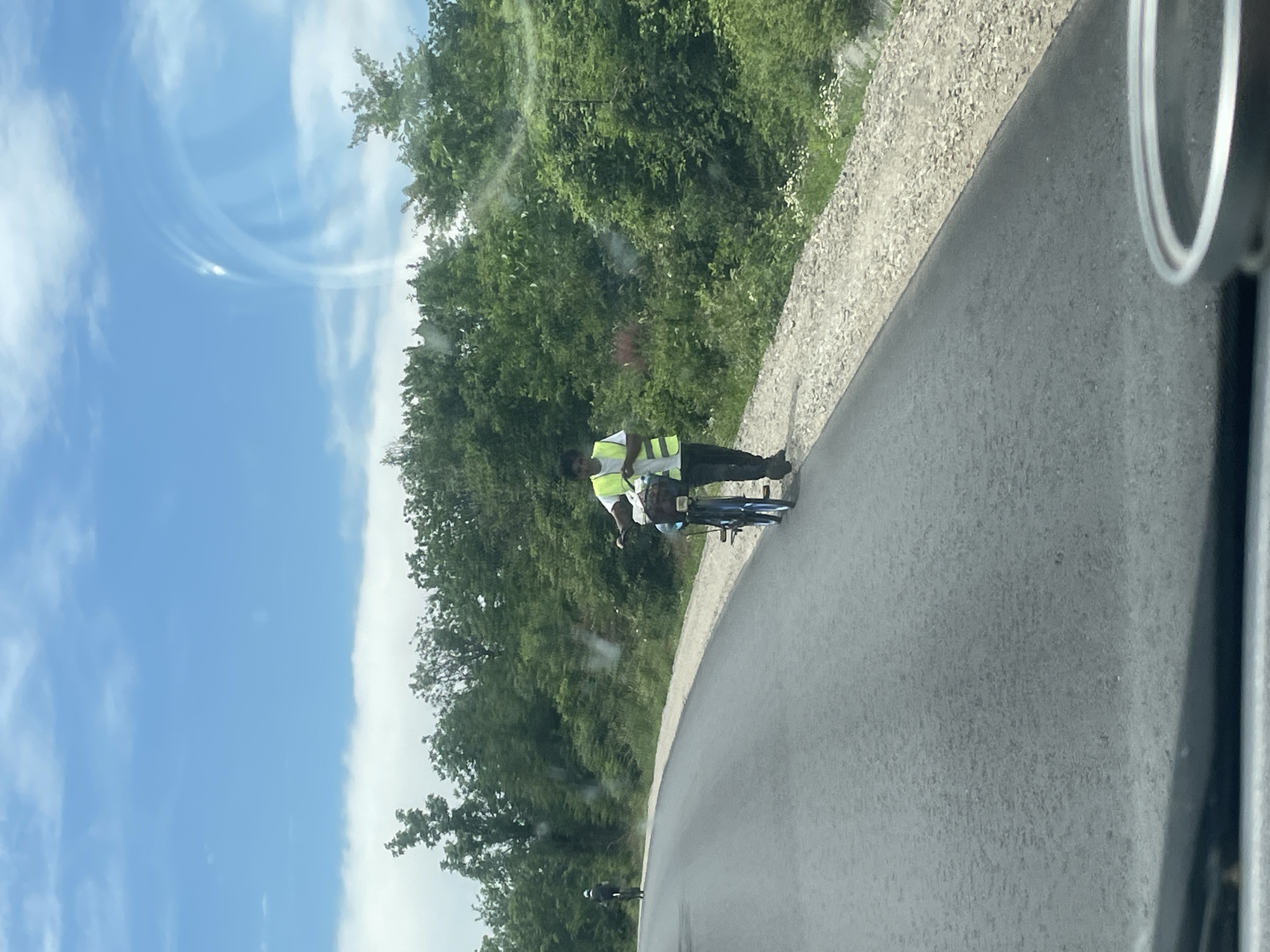 The image shows a man in a yellow vest standing next to a blue bicycle on a paved road, possibly on the outskirts of Probuda, Bulgaria. The road curves to the right, and on the left side of the frame is a line of trees and vegetation, followed by a blue sky with white clouds. On the right, there's a gravelly area next to the road. The photo appears to be taken from inside a moving vehicle.