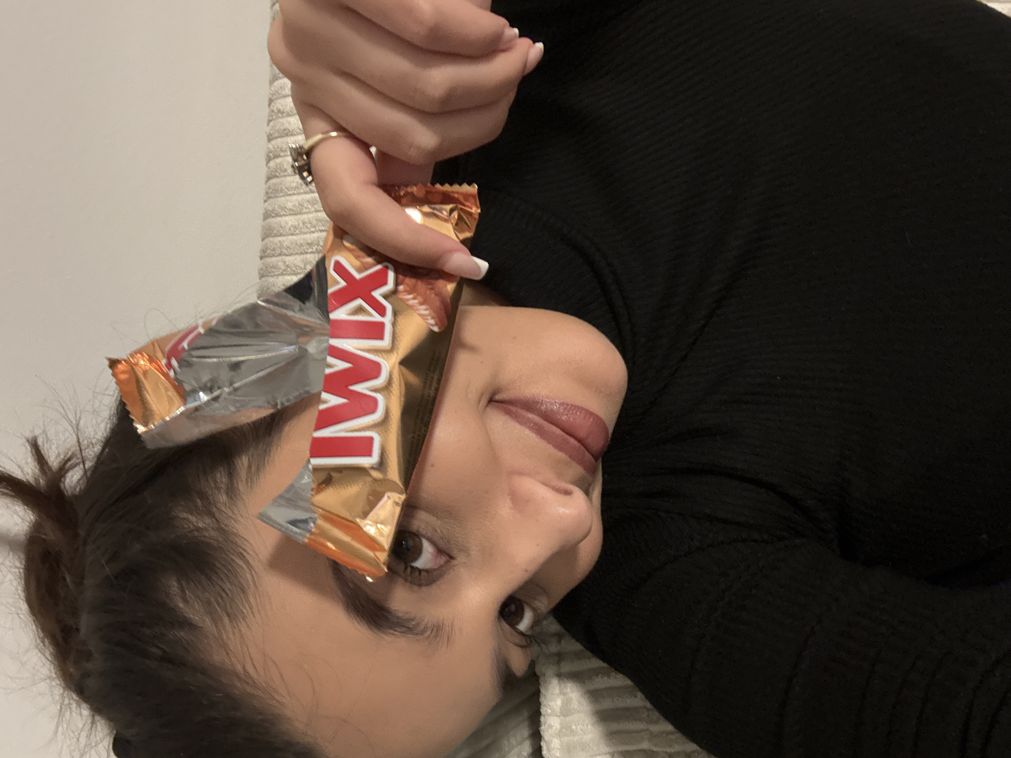 A person is holding a Twix candy bar in front of their face. The candy bar covers part of the person's forehead and eye. The candy bar's wrapper is gold and red and the word "TWIX" is printed on it. The person has brown eyes, dark hair, and is wearing a black shirt. The background is a neutral color. The city of Mönchengladbach, Germany is not discernible in this image.