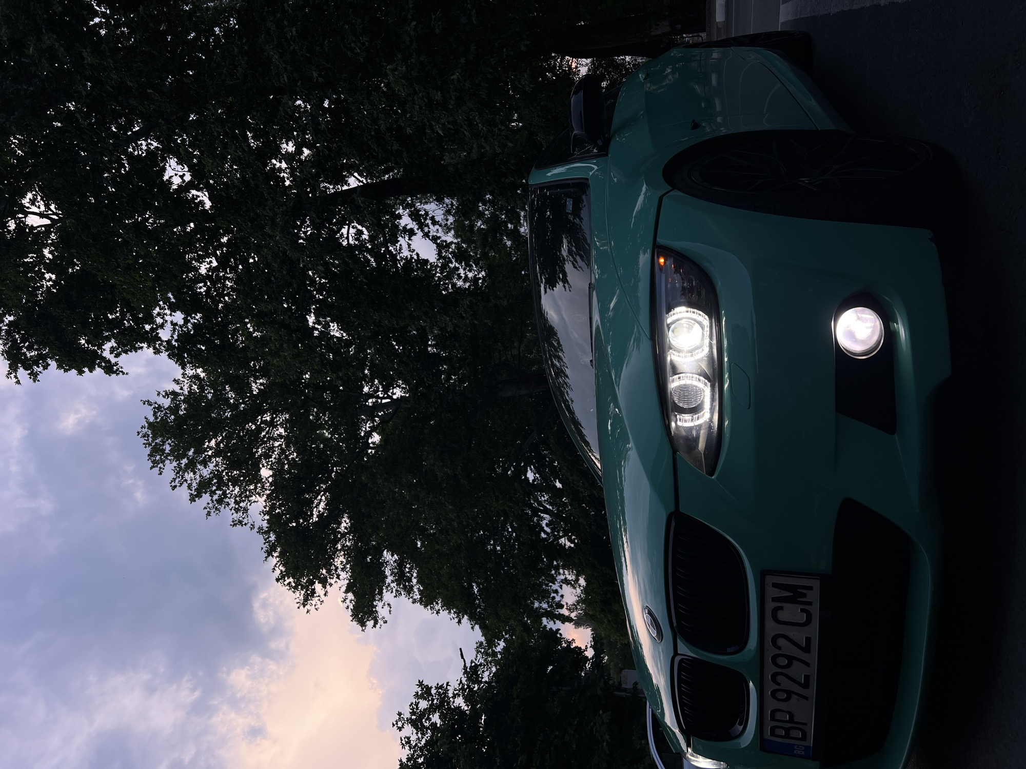 An exterior, low-angle photograph captured at dusk or twilight in Asenovgrad, Bulgaria, features a teal-colored BMW 3 Series coupe (E92 generation) parked outdoors. The vehicle, positioned in the foreground with its front-left quarter prominent, displays illuminated LED 'angel eye' daytime running lights and a lit front-left fog light. Its front grille is black, and a Bulgarian license plate, 'BP 9292 CM', is affixed to the bumper. The car is equipped with dark-colored alloy wheels. To the immediate left of the BMW, a portion of a light-colored vehicle is partially visible. The background is dominated by a dense canopy of large, dark-leaved trees, silhouetted against a cloudy sky exhibiting twilight hues of purple, pink, and orange near the horizon, transitioning to a lighter grey-blue higher up. No individuals are present in the scene. A white parking space line is discernible on the dark asphalt surface in the lower-right foreground.