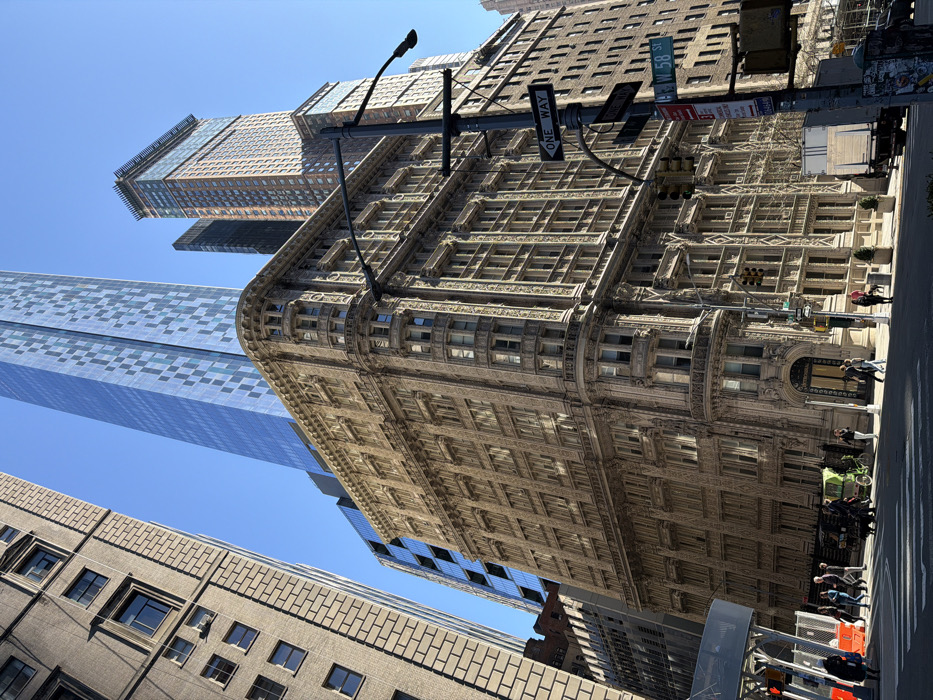 The image depicts an urban street scene in Midtown Manhattan, New York City, United States of America, captured on a clear, sunny day with a bright blue sky and prominent shadows. The perspective looks generally north from near the intersection of W 58th Street and Broadway.
In the midground, a multi-story building known as the Gainsborough Studios, featuring a light-colored, highly ornate stone facade with intricate carvings, pilasters, and decorative window frames, stands at a rounded corner. To its immediate left, a taller, plainer building with a brick or stone facade is visible. Dominating the background skyline is a very tall, modern skyscraper with a reflective blue glass facade patterned with lighter squares, identified as Central Park Tower. To the right of the Gainsborough Studios, other tall buildings with varying facades extend into the distance.
The paved street in the foreground features marked crosswalks. A dark gray street light pole, equipped with multiple arms, holds traffic lights and street signs; one sign clearly reads "W 58 ST" and another "ONE WAY." Approximately 10 to 15 pedestrians are traversing the crosswalk from left to right, some individually, some in small groups, with one person walking a dog. Additional pedestrians are visible on the sidewalks. A lime green bicycle is positioned near the left side of the crosswalk. A white box truck is stopped or parked on the right side of the street, partially obscured by the street pole. Potted plants or small trees line the sidewalk near the ornate building's entrance. A white scaffolding structure is present on the far left side of the scene.